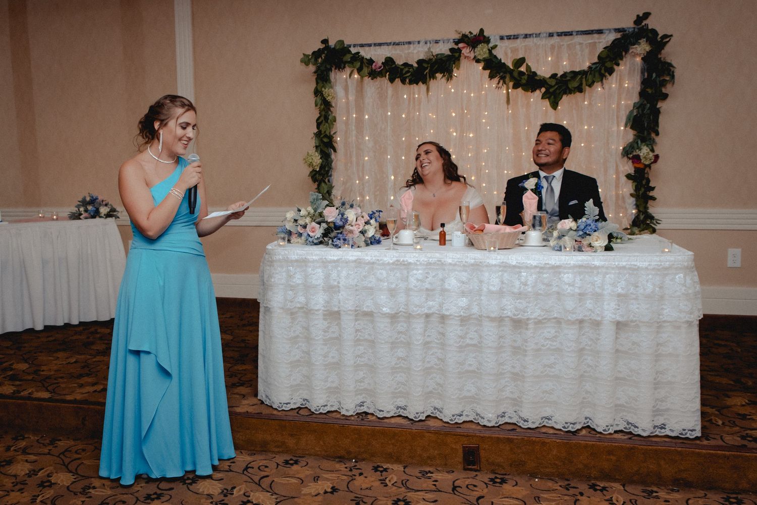 The maid-of-honor in a flowing blue dress shares a toast as the bride & groom sit at the head table laughing and smiling in Westchester NY.