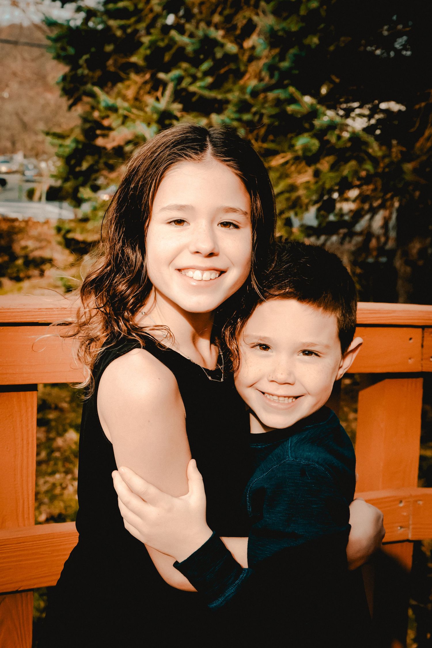 Siblings hug while smiling at the camera. A wood bridge overlooking evergreen trees in Westchester, New York is in the background.