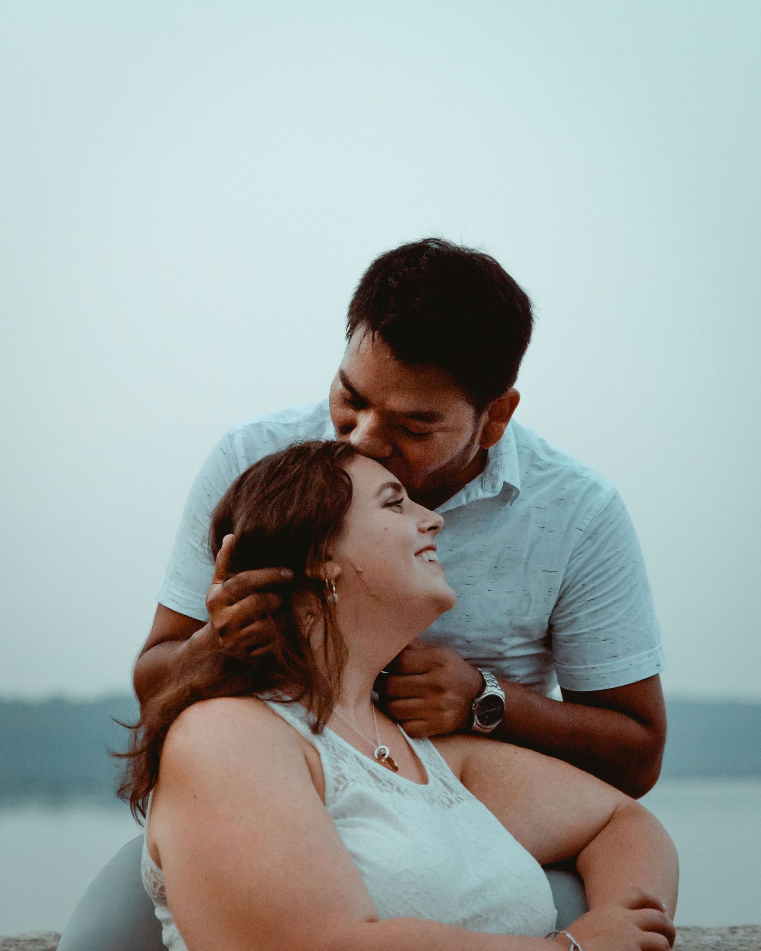 A couple shares a romantic moment at dusk atop the Kensico Dam in Valhalla, NY. The man is kissing his fiancee's forehead.