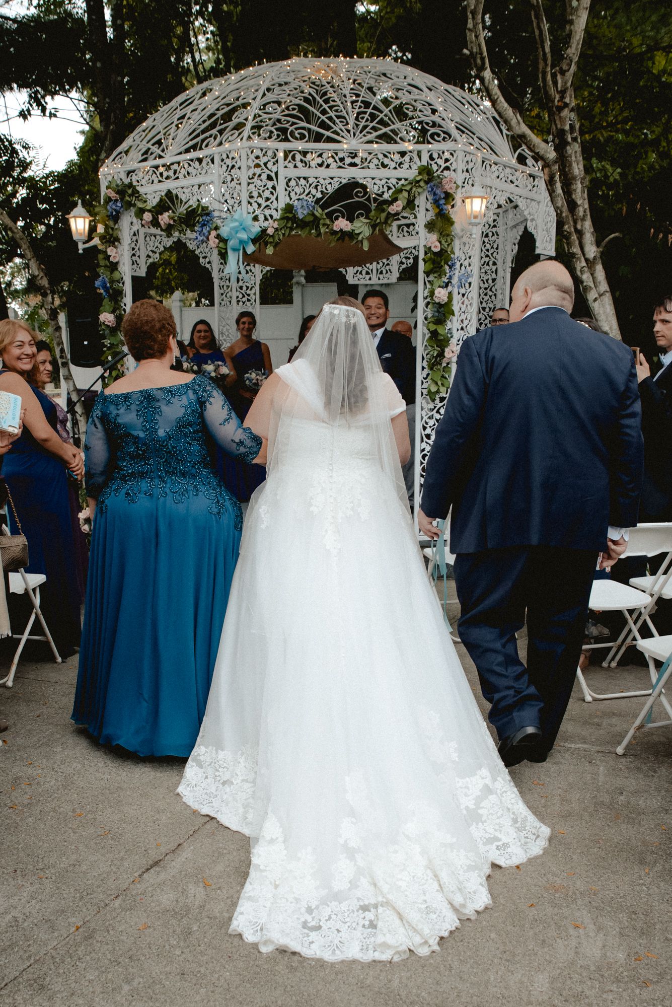 A bride being walked down the aisle by her mom and dad arm-in-arm. Photographed from behind, her long flowing train is front and center.