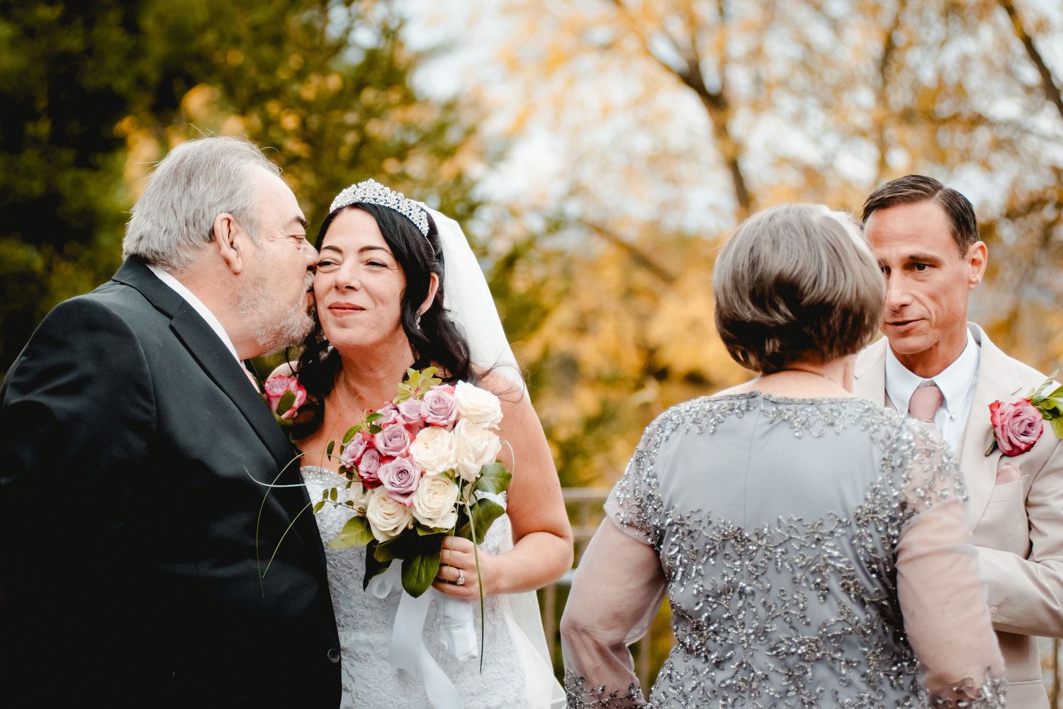Father of the bride kisses her on the cheek while the groom and his mother embrace next to them, smiling. Fall leaves set the scene in NY.