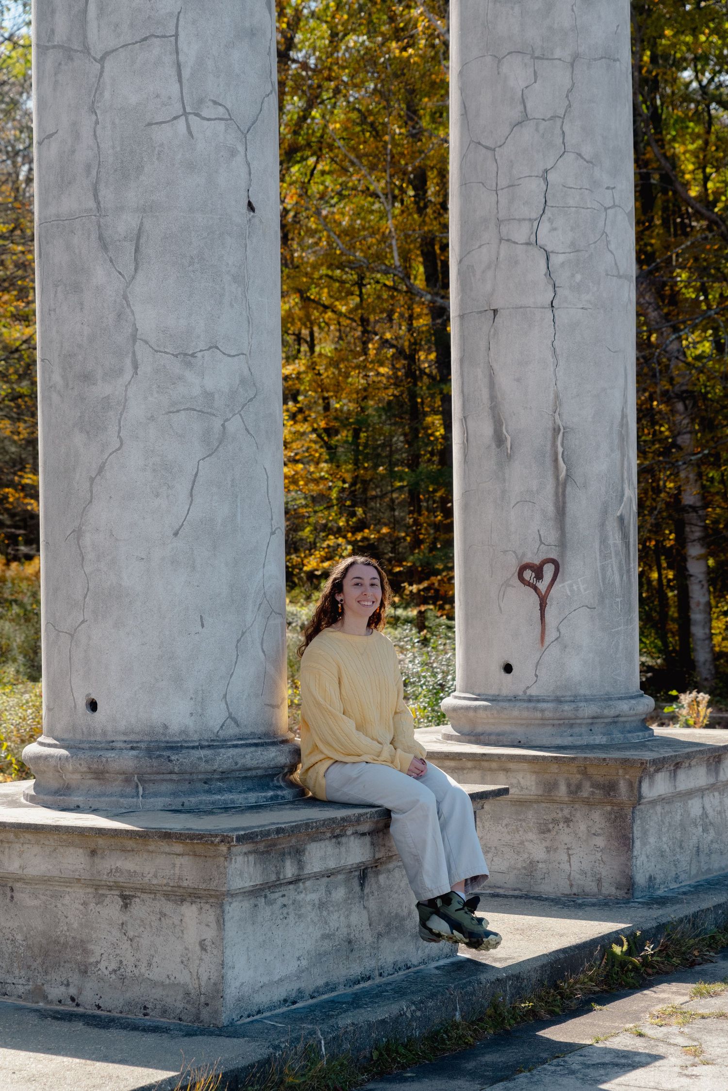 The wedding photographer wears a yellow sweater while sitting between two tall columns, one of which has a heart drawn on, on a fall day.