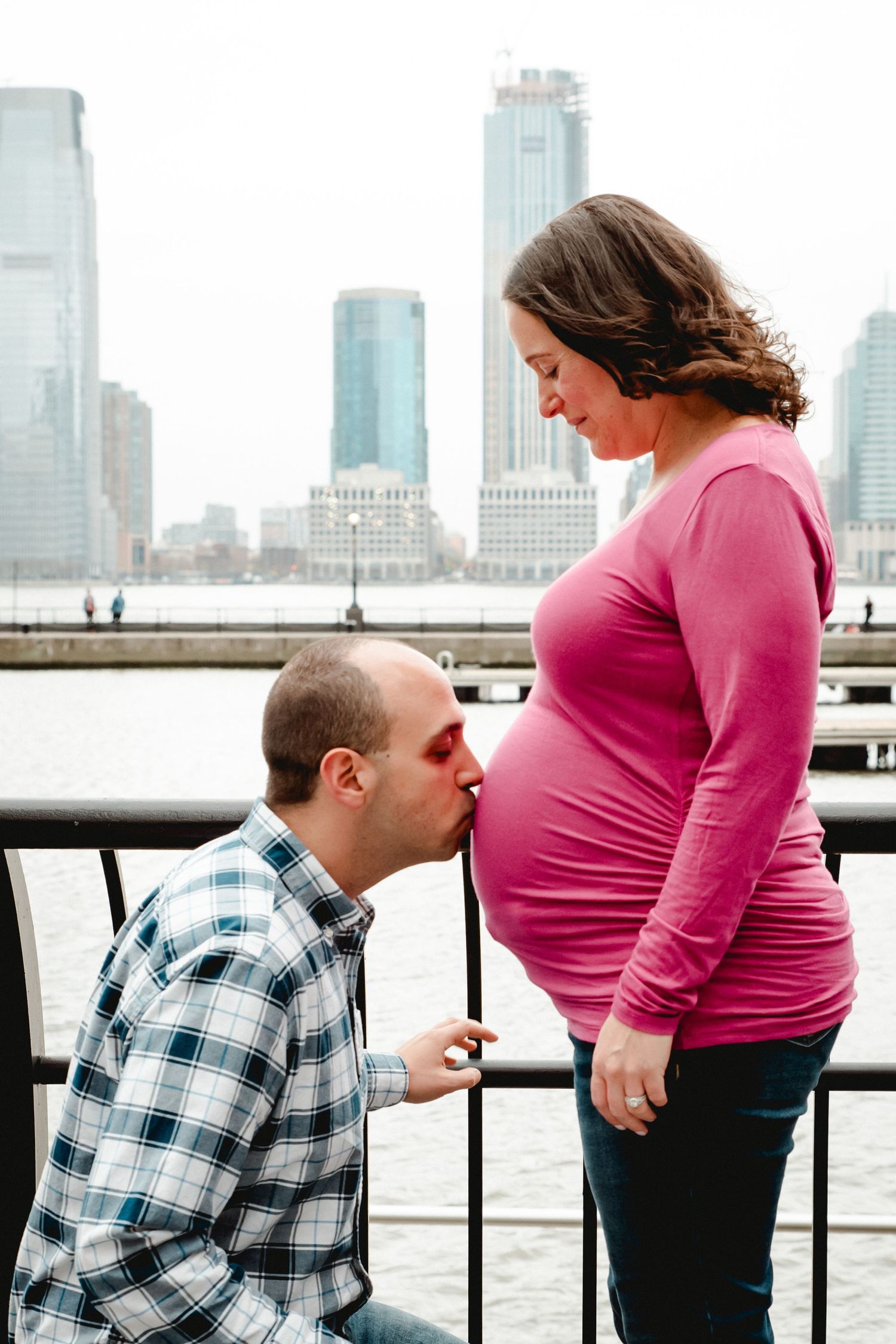 Overlooking the river and skyline, a man kisses his wife's baby-bump while she looks down at him smiling.