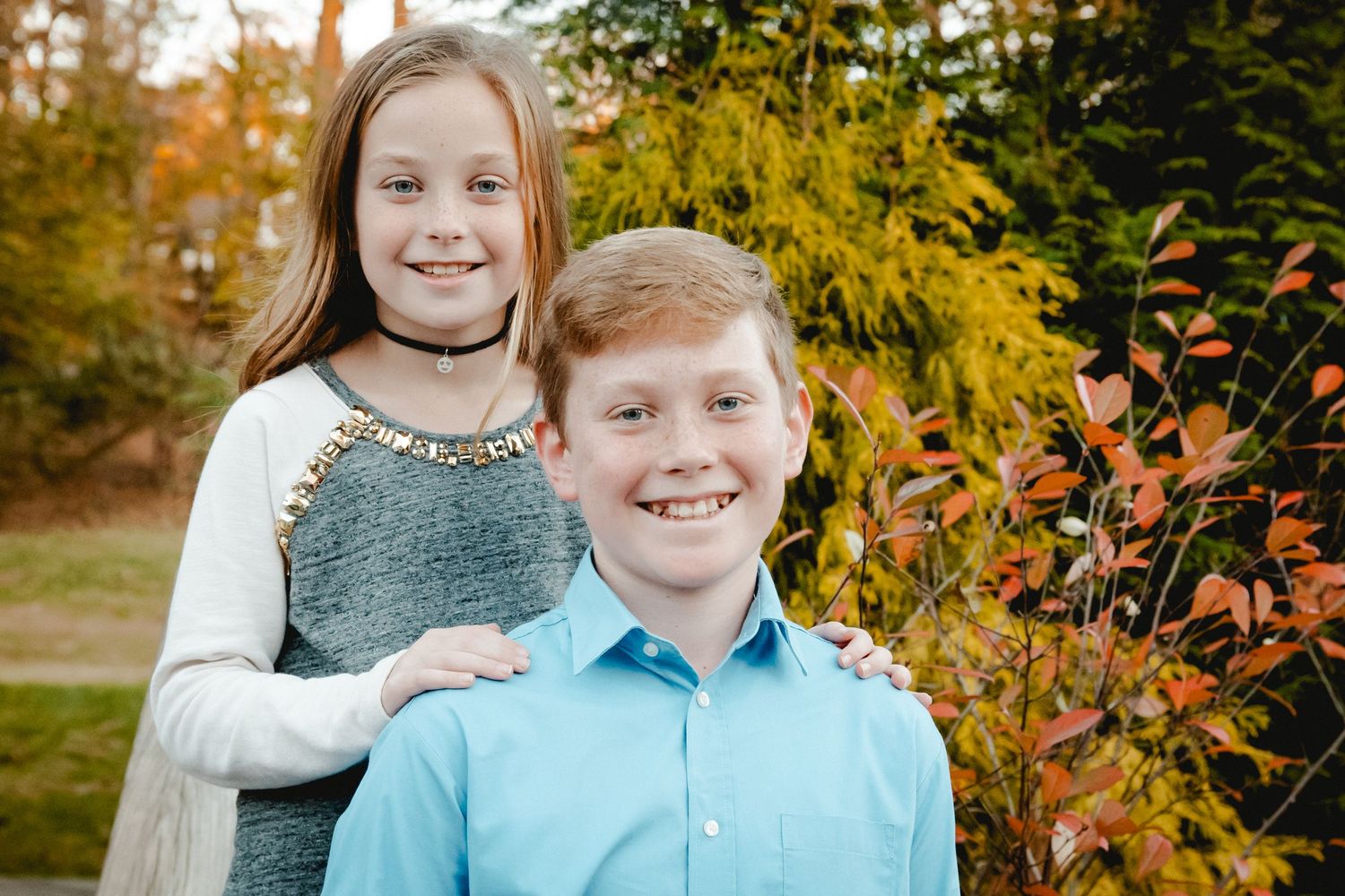 A boy and girl smiling at the camera on a stunning fall afternoon in Westchester, NY. The girl's hands are on her big brother's shoulders.