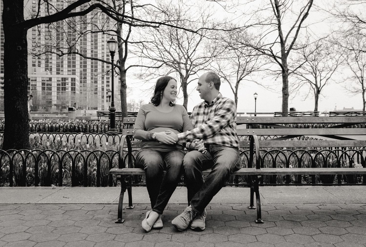 An NYC-based couple sits on a quiet park bench in NYC. The woman is pregnant and her husband holds her baby-bump while smiling.