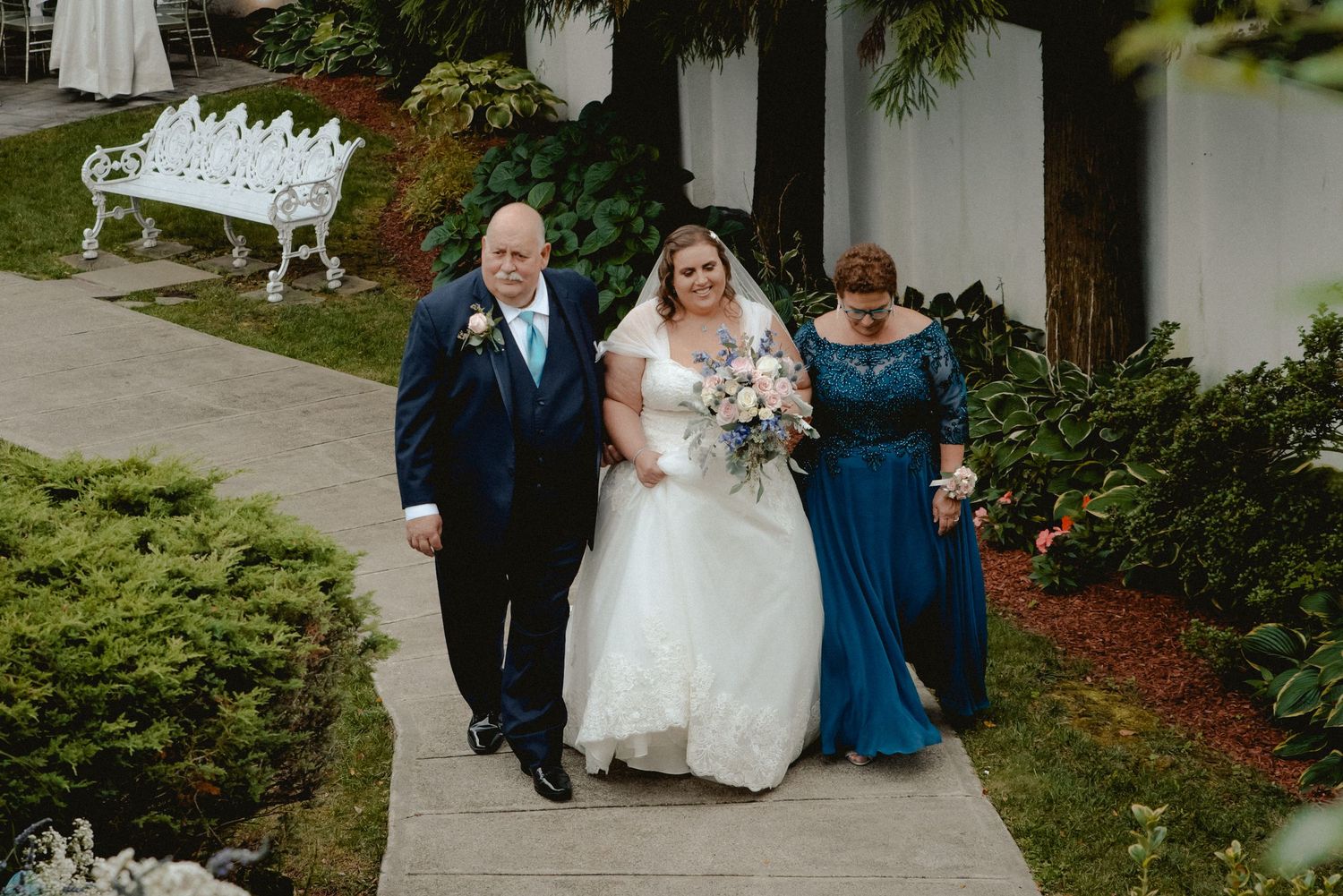 A smiling bride holding a beautiful pink bouquet links arms with her parents to begin her walk down the aisle to marry her groom.