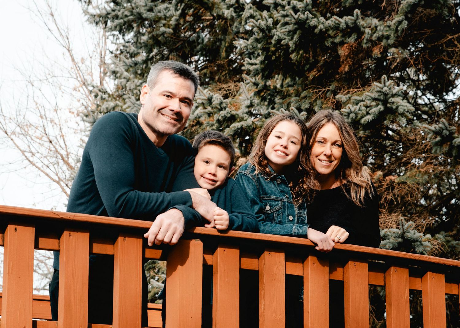 A family of four stands atop a scenic wooden bridge. Everyone is standing close together and smiling. There are lush trees behind them.