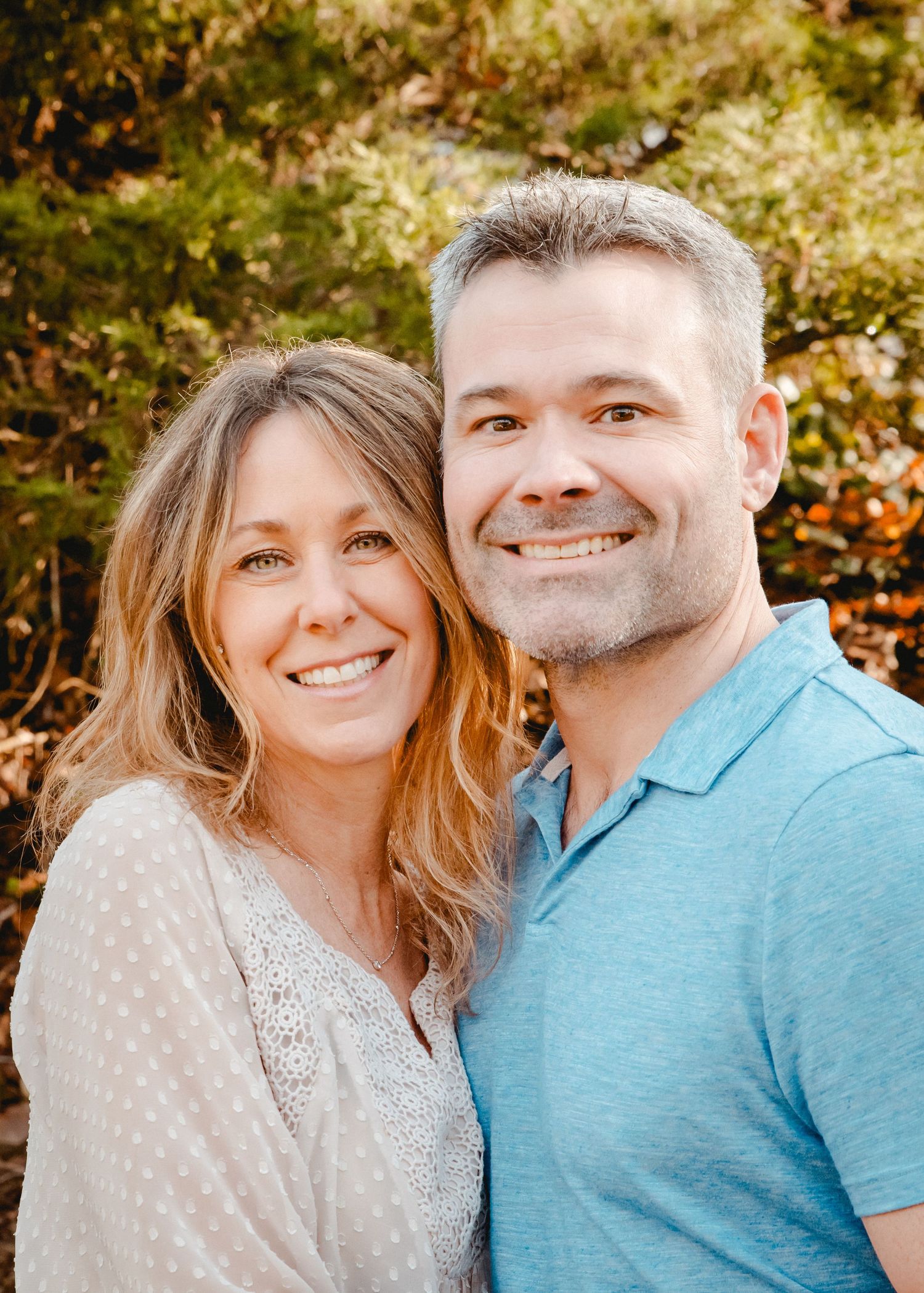 Husband and wife smiling at the camera for a casual portrait in their Westchester, NY backyard surrounded by greenery.