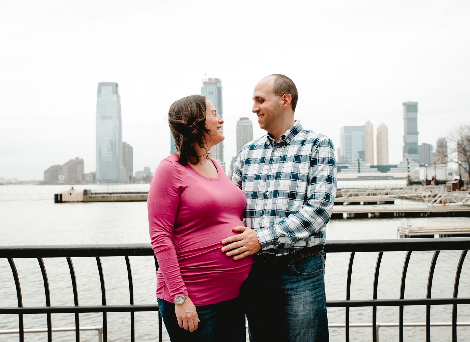 A pregnant woman looks up at her husband smiling while he holds her baby-bump. The couple overlooks the river in downtown New York City.