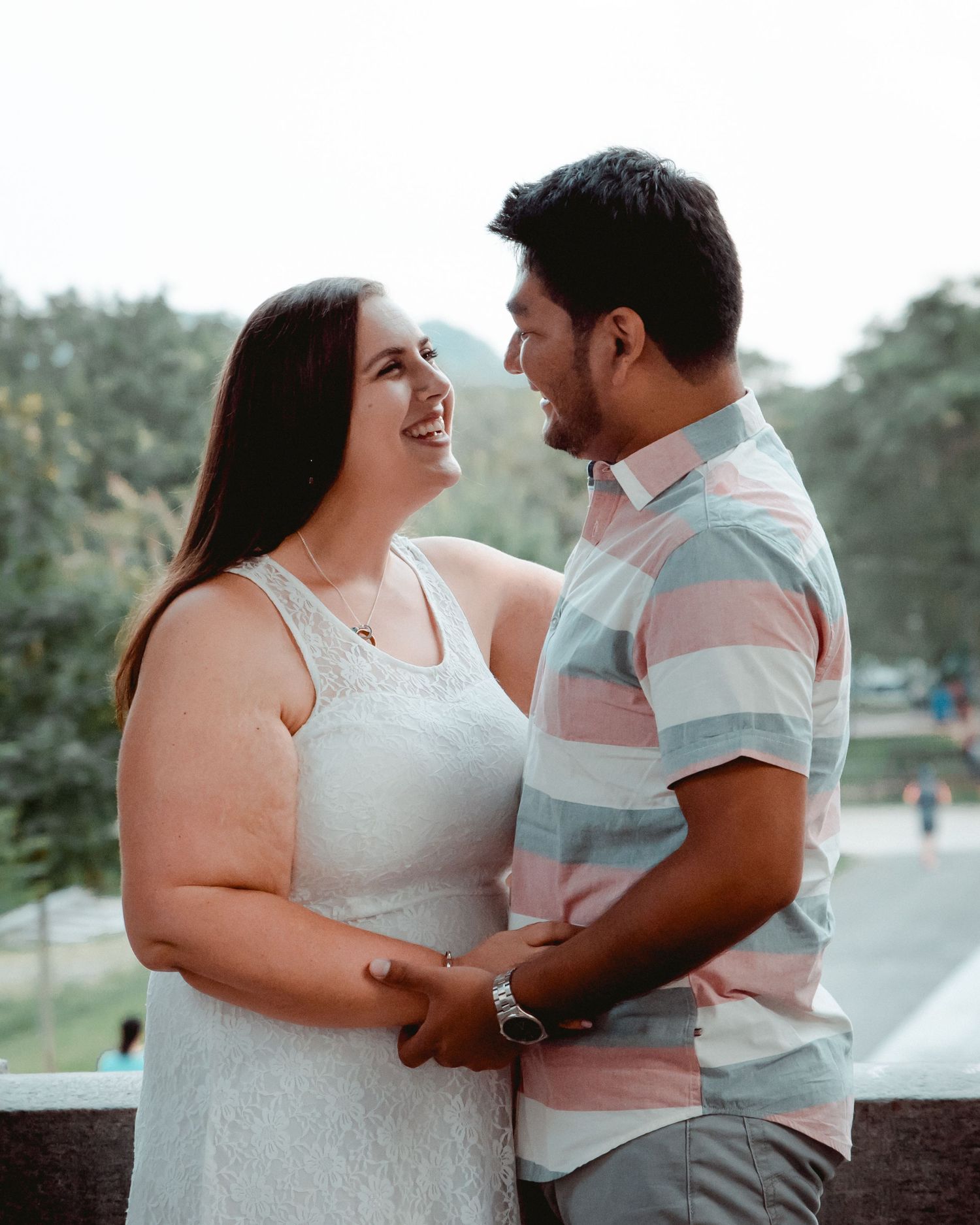 A newly engaged couple share an embrace and look into each other's eyes while smiling. Soft, lush greenery in the background.