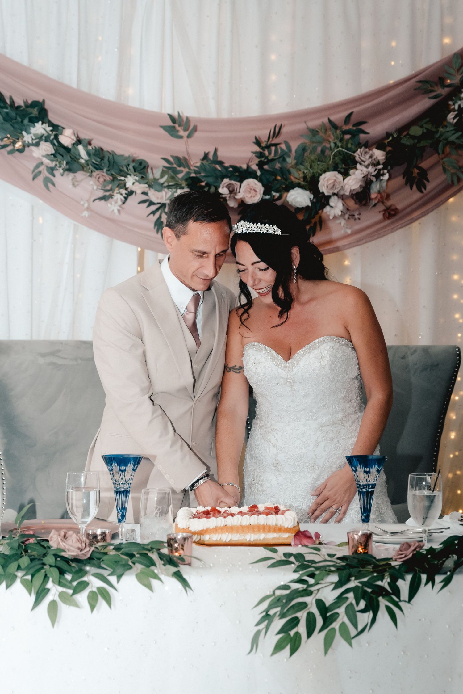 The bride and groom cutting their wedding cake at the head table at the Riverdale in Endicott, NY. They are surrounded by flowers.