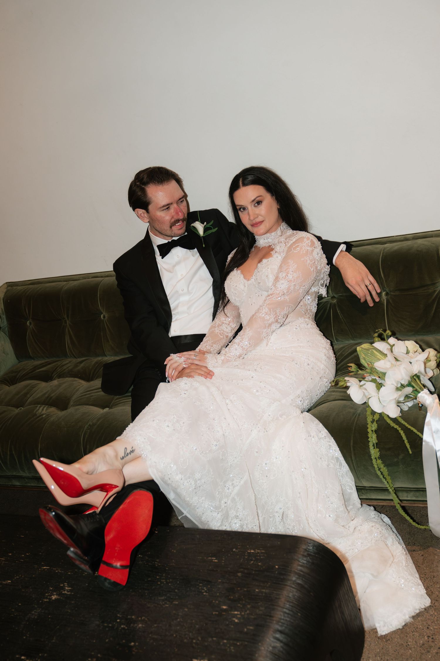 Bride in lace gown with red Louboutins sits with groom in black tuxedo on green velvet sofa.