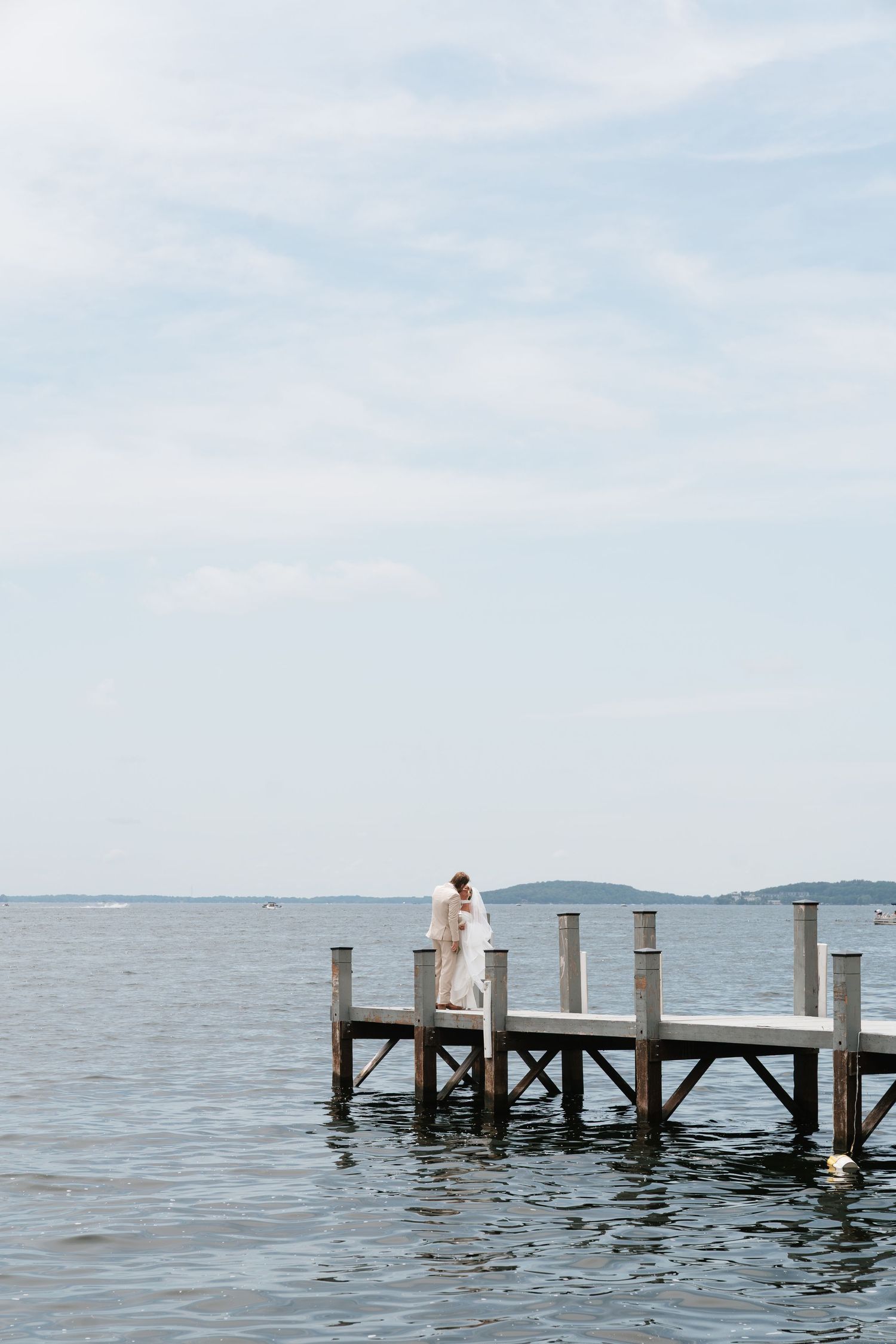 Couple embracing on a wooden dock over calm water under a partly cloudy sky, with distant shoreline in the background.
