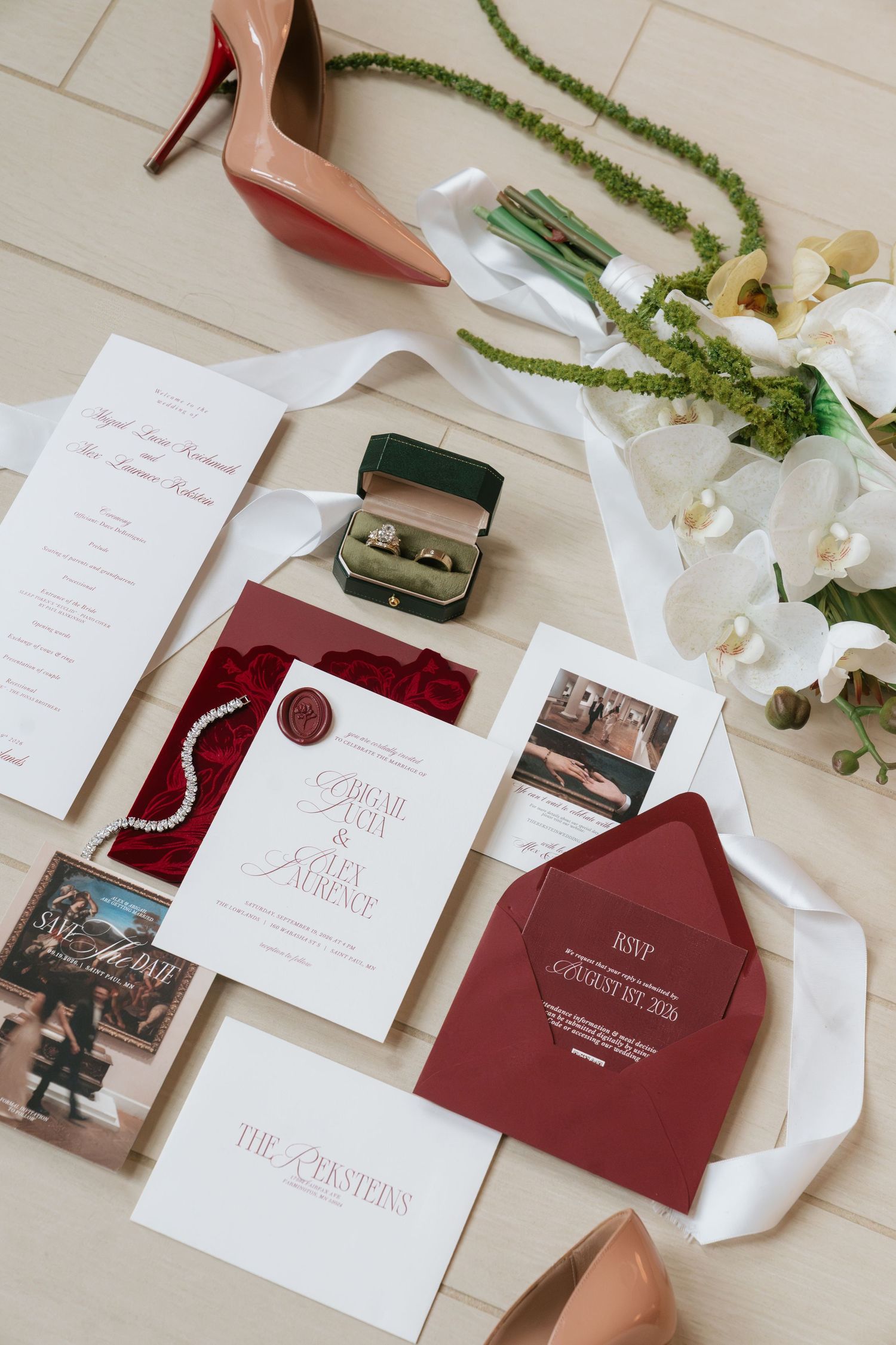 Elegant wedding flat lay with burgundy invitations, diamond bracelet, ring box, heels, and white orchid bouquet on light floor.