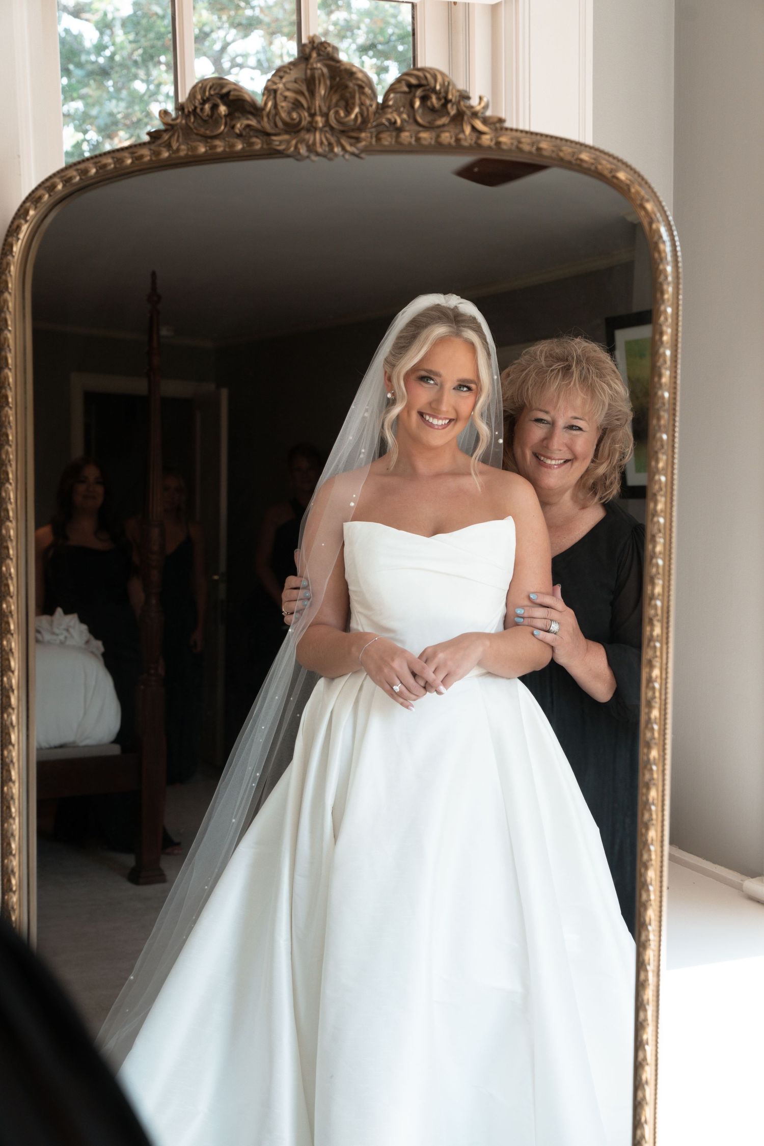 A reflection in an ornate gold mirror shows a bride in a white strapless gown standing with her mother. Hilltop House, Rochester, MN.