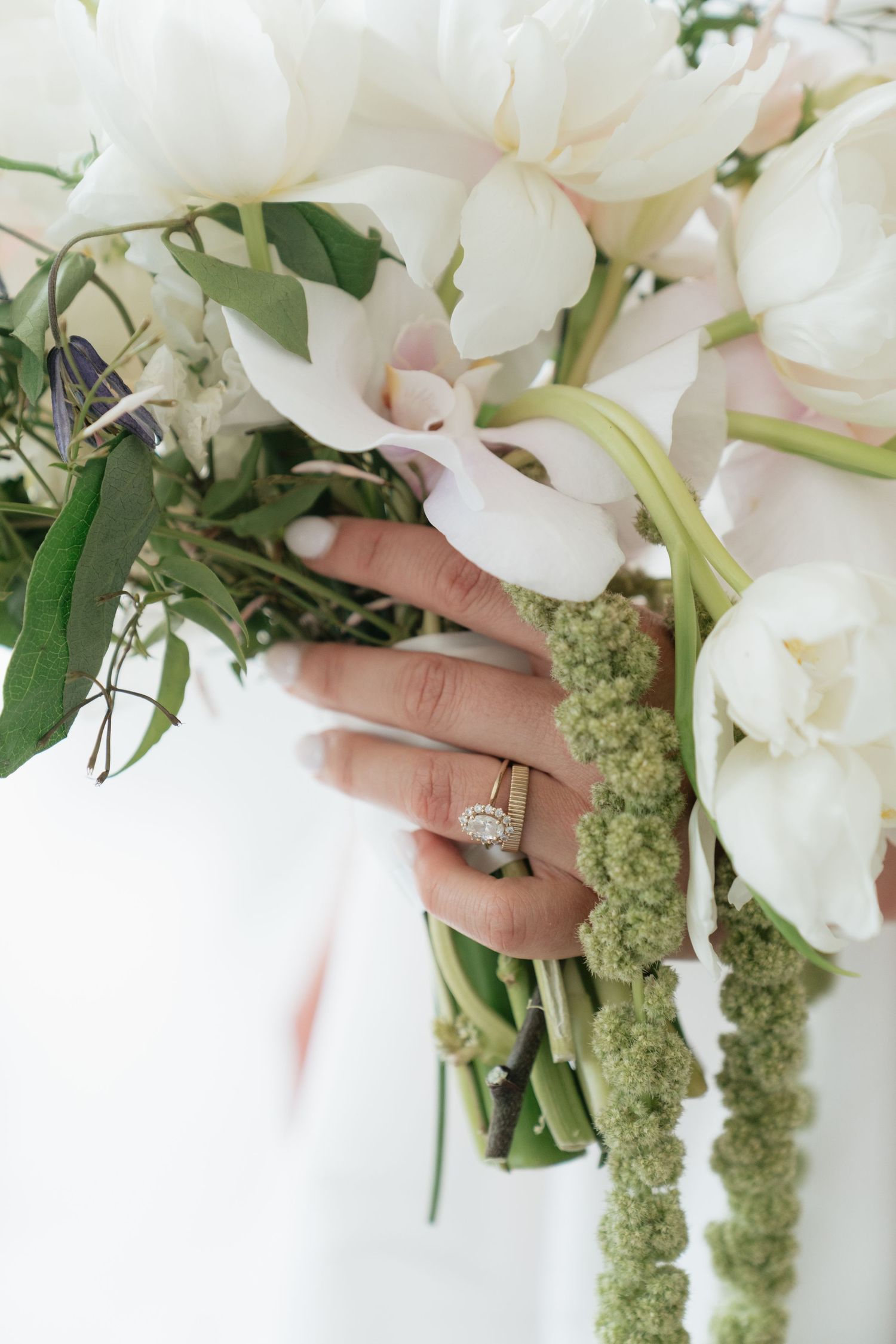 Bride holding white orchid bouquet with diamond engagement ring and gold wedding band on manicured hand.