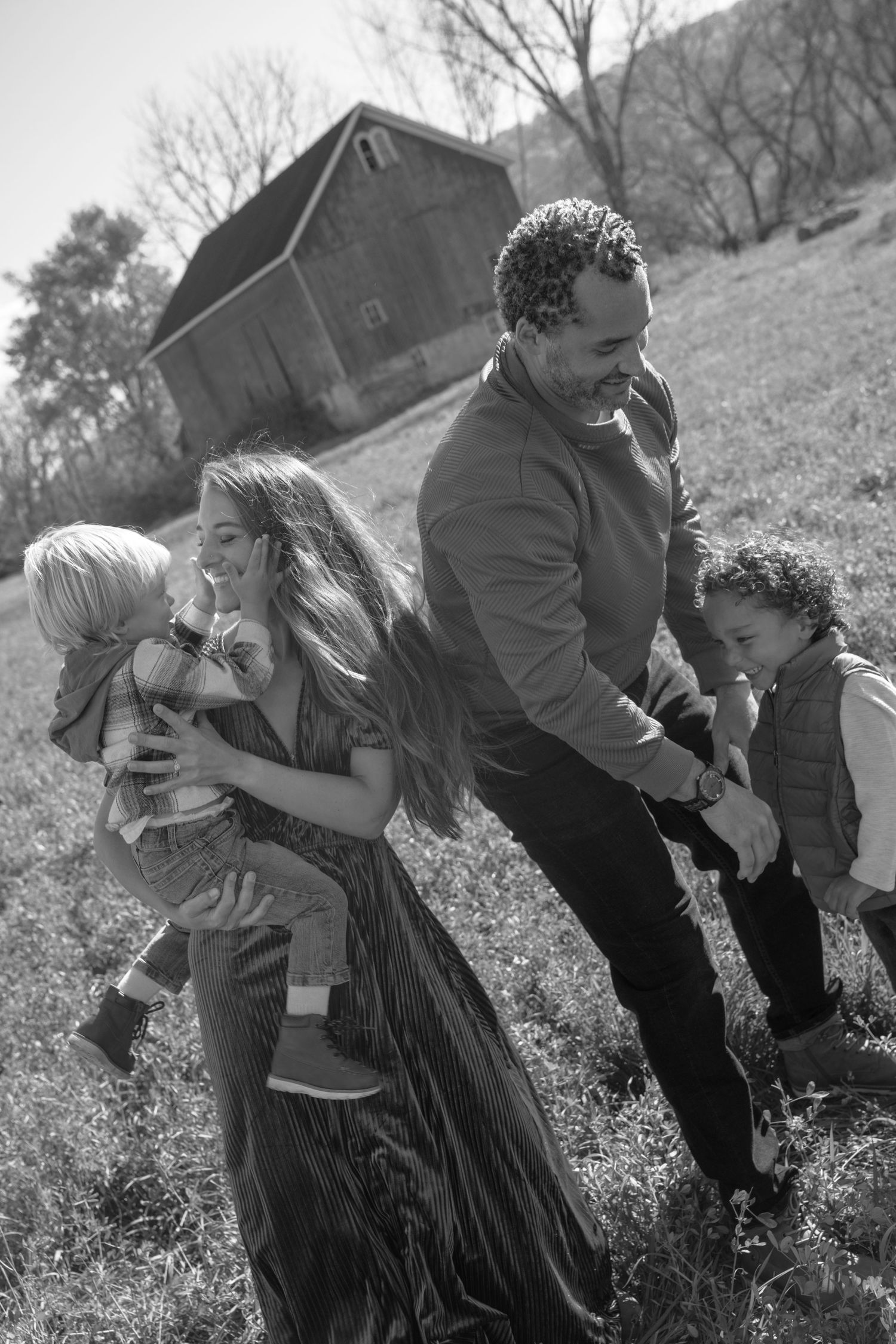 A family enjoying a playful moment together on a grassy hillside with a rustic barn in the background.