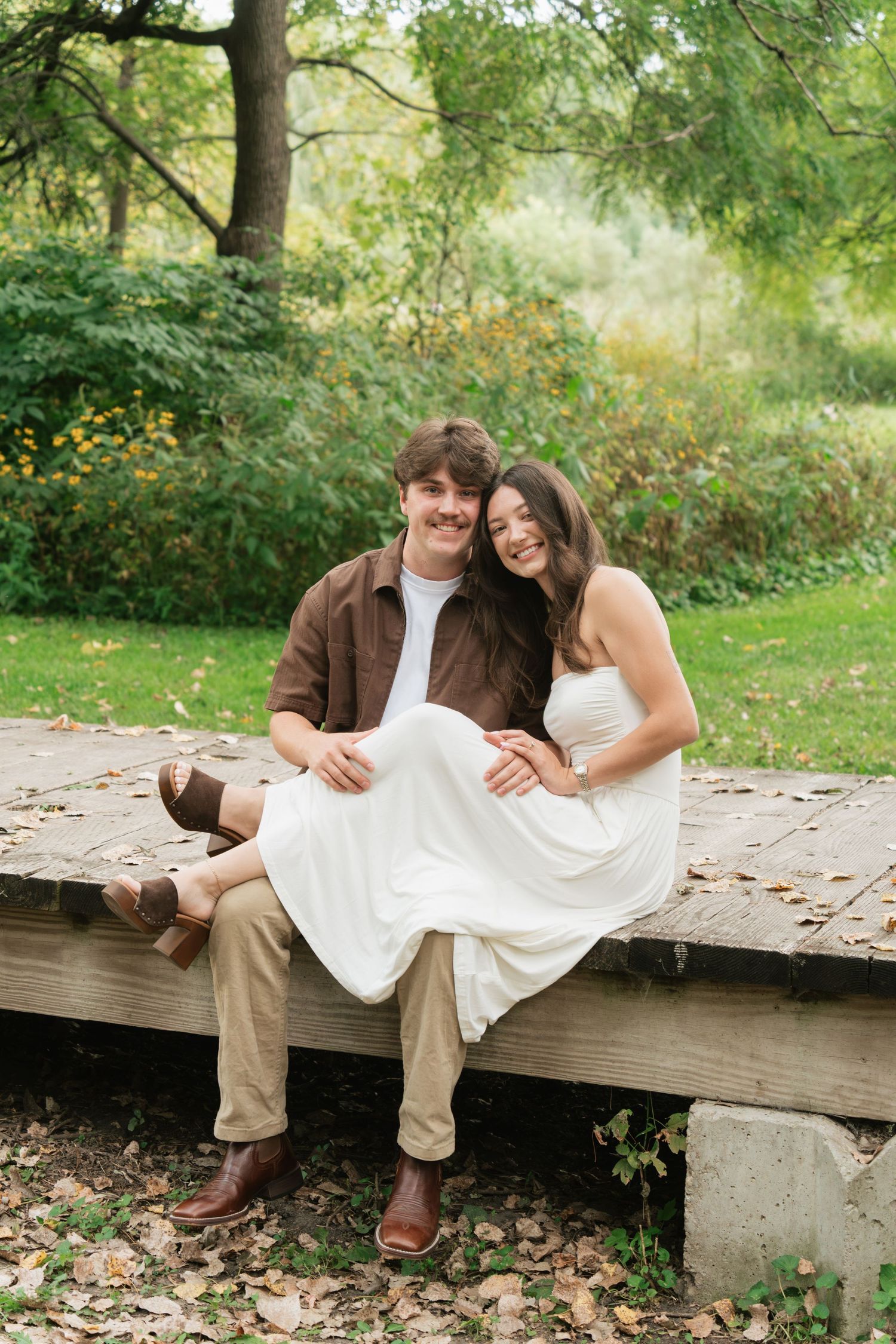 A couple sits on a wooden platform in a lush park, smiling, man in brown jacket and woman in white dress.