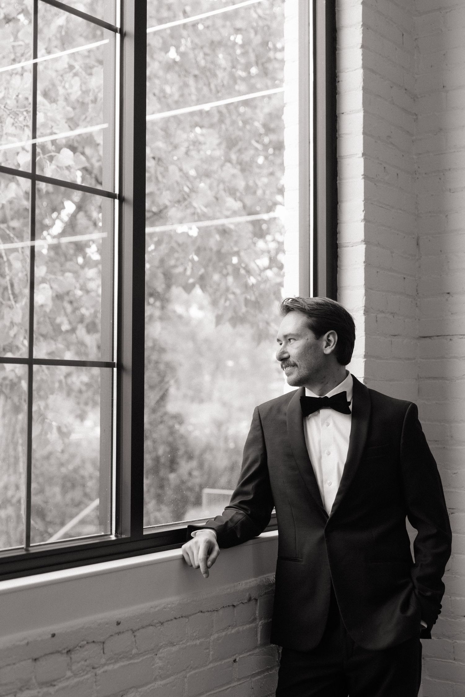 Elegant groom in black tuxedo and bow tie standing by large window in black and white wedding photo.