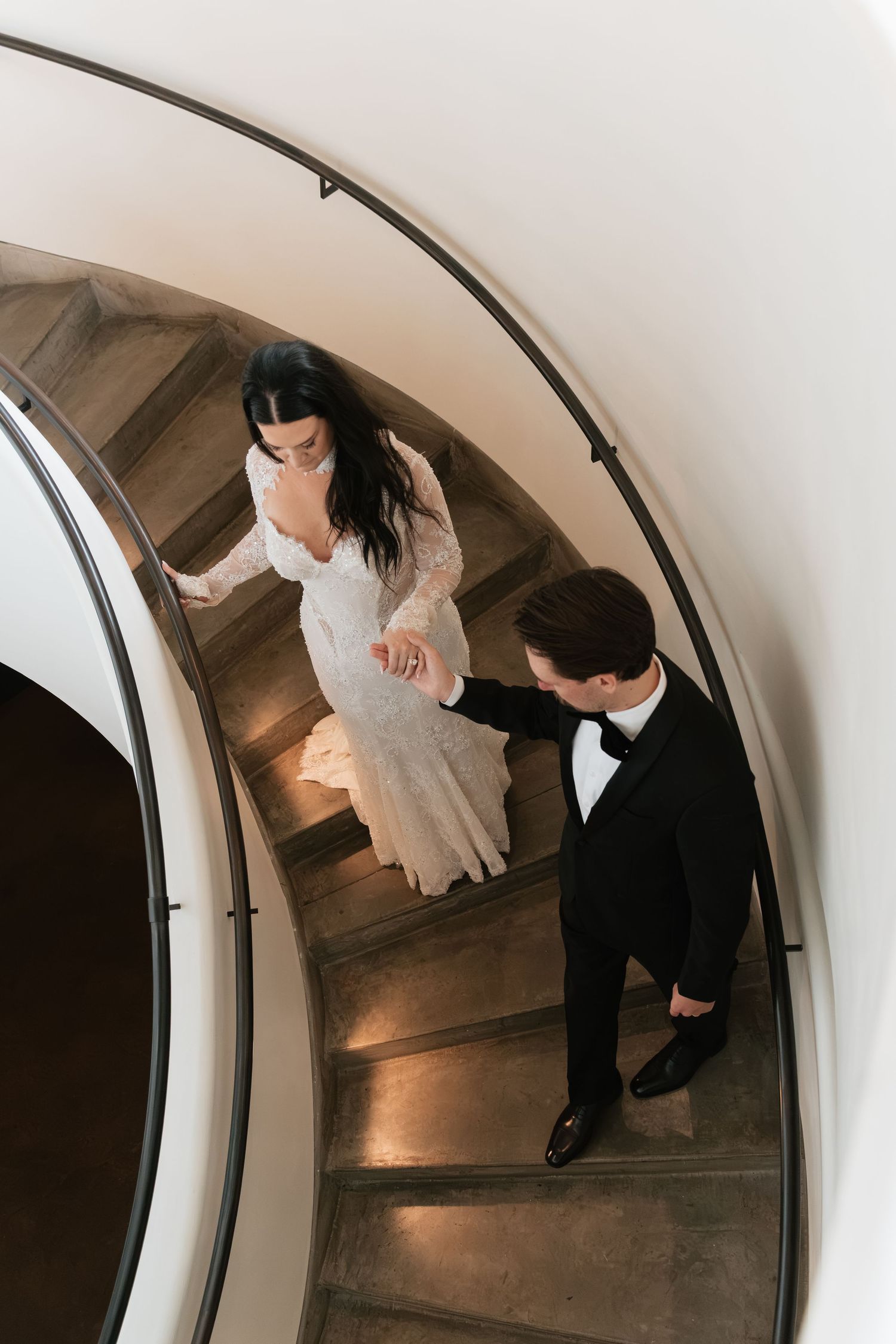 Aerial view of bride in lace gown and groom in tuxedo holding hands on a curved spiral staircase.