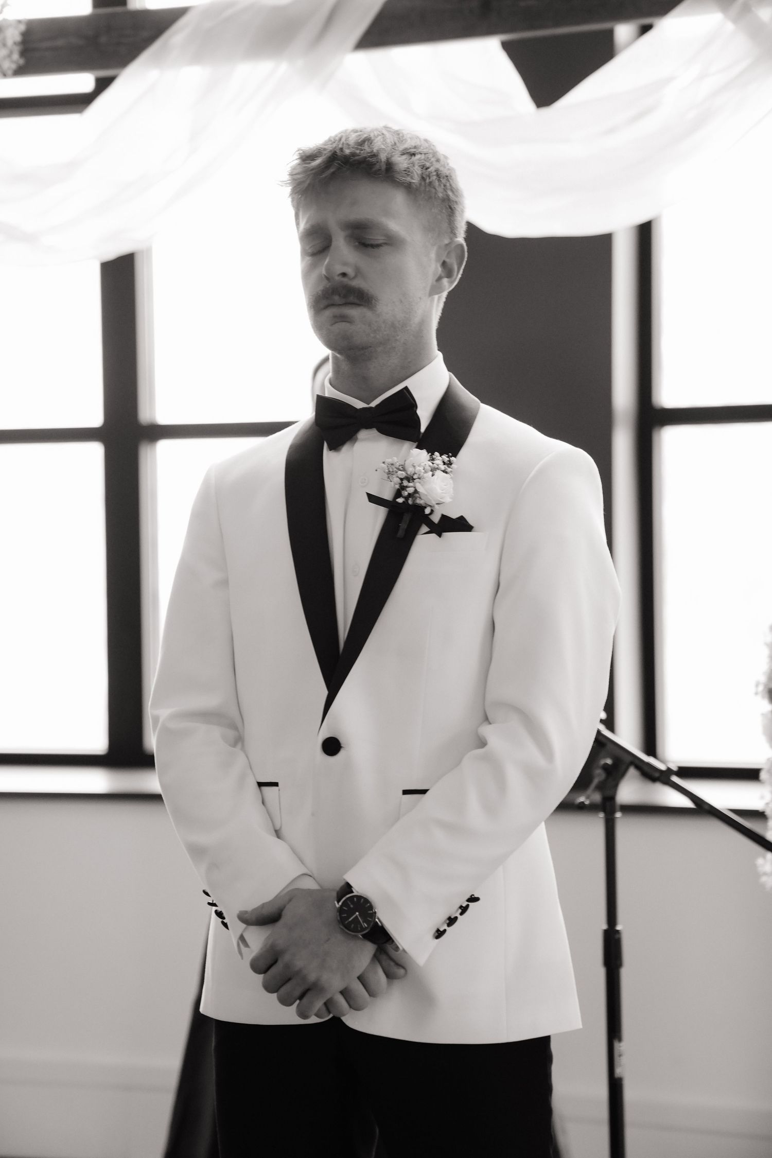 Black and white photo of a groom in white tuxedo jacket with bow tie and boutonniere, eyes closed at wedding ceremony.