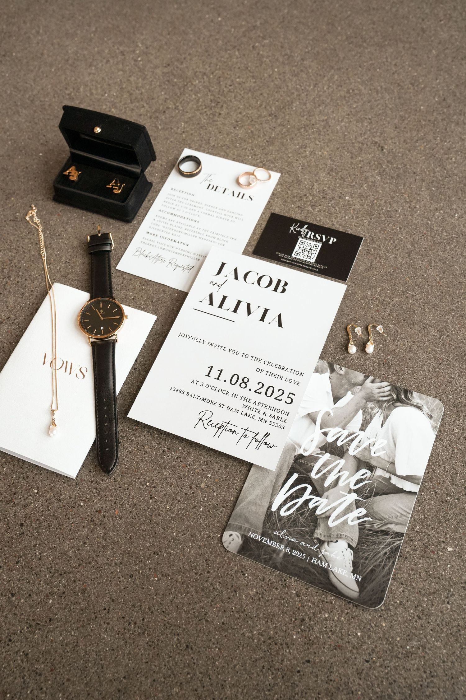 Elegant wedding flat lay featuring save the date cards, vows booklet, black watch, gold jewelry, and RSVP cards on gray surface.
