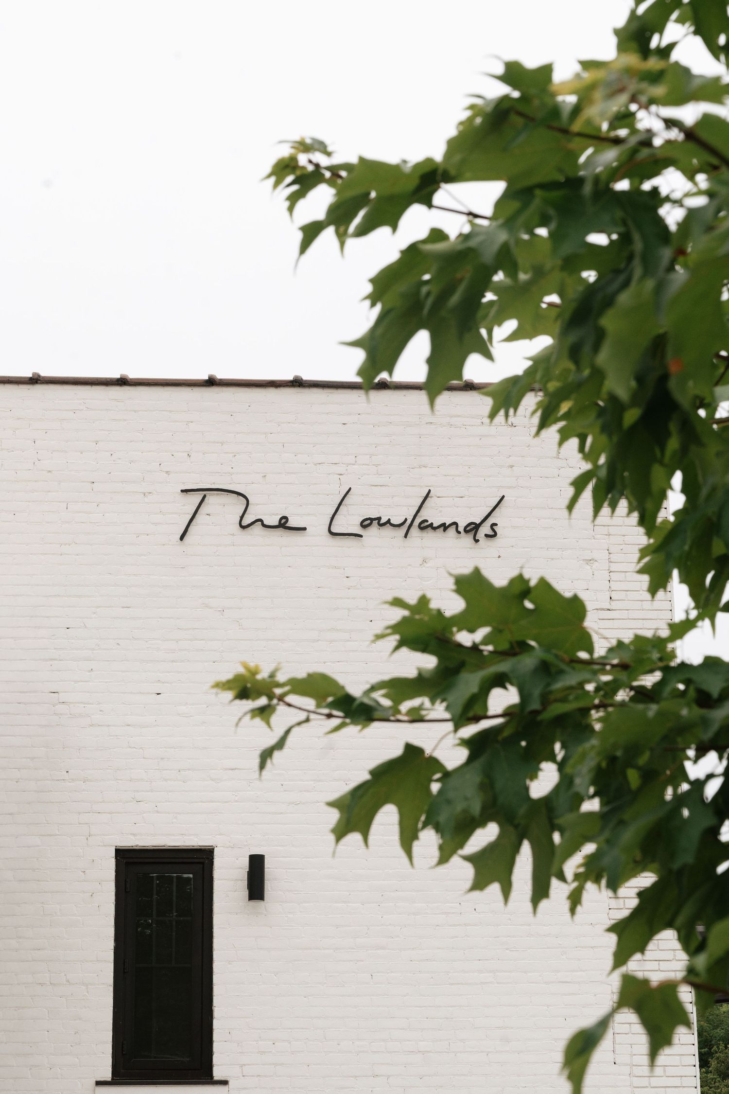 White brick exterior of The Lowlands venue with cursive signage and green maple tree branches in foreground.