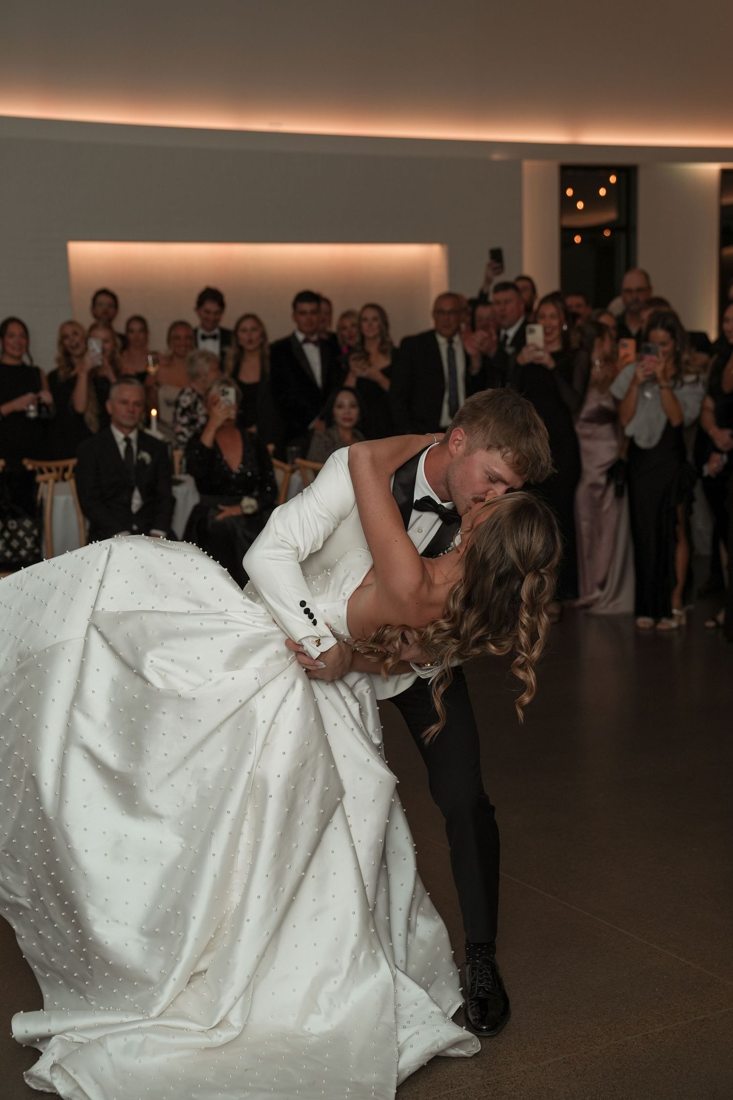 A couple performs a dramatic dip during their first dance at a wedding reception while guests watch in the background.