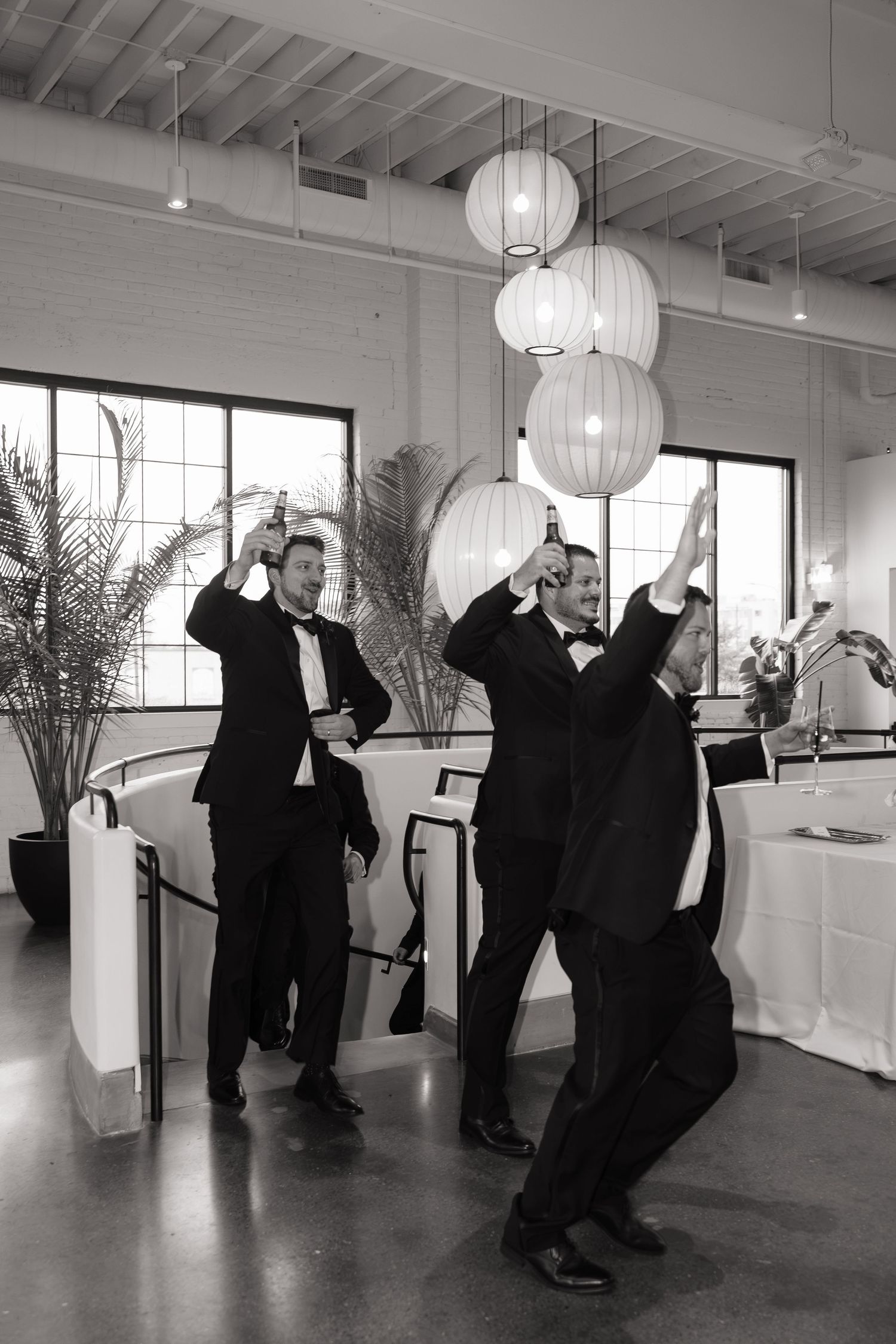Groomsmen in tuxedos celebrate joyfully at a wedding reception in a modern venue with globe pendant lights.