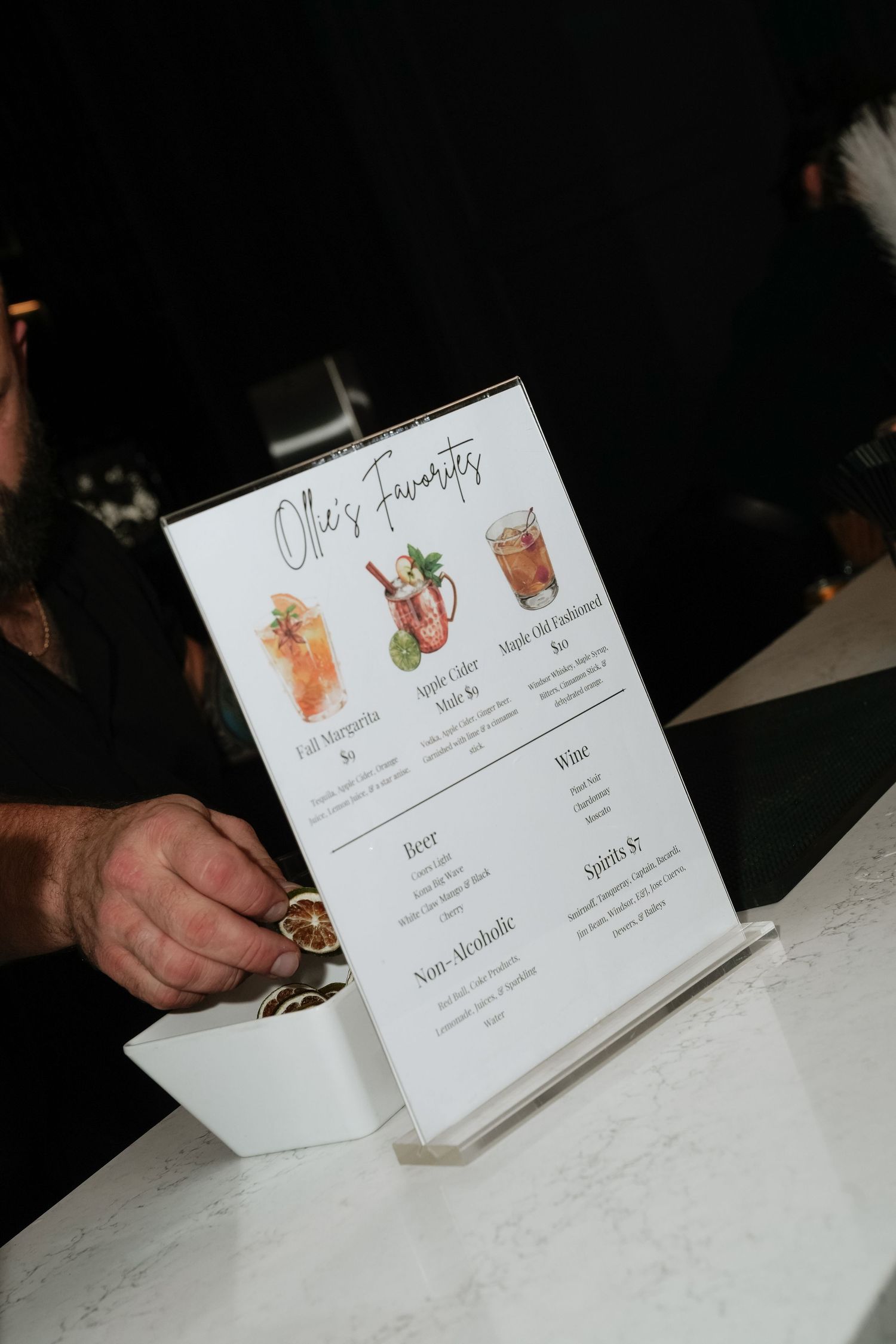 A hand holds a cocktail menu displaying watercolor illustrations of drinks on a white marble countertop.