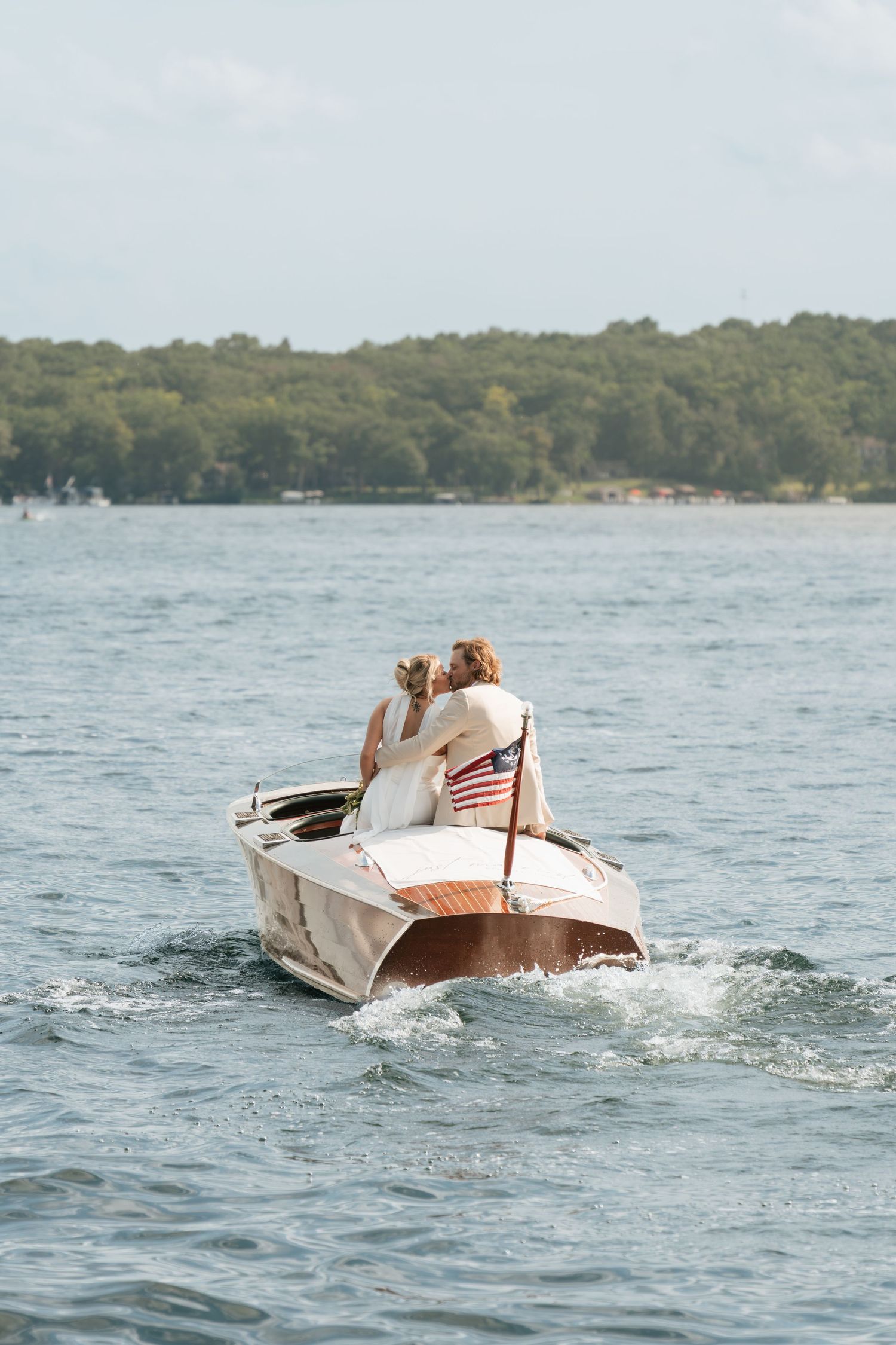 Two people ride in a classic wooden motorboat with an American flag on a lake surrounded by trees.