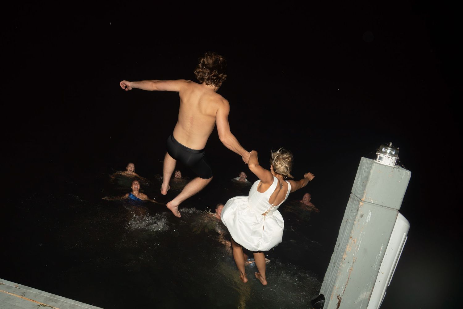 Young adults jumping into dark water at night, with a shirtless man and woman in white dress leaping from a dock.