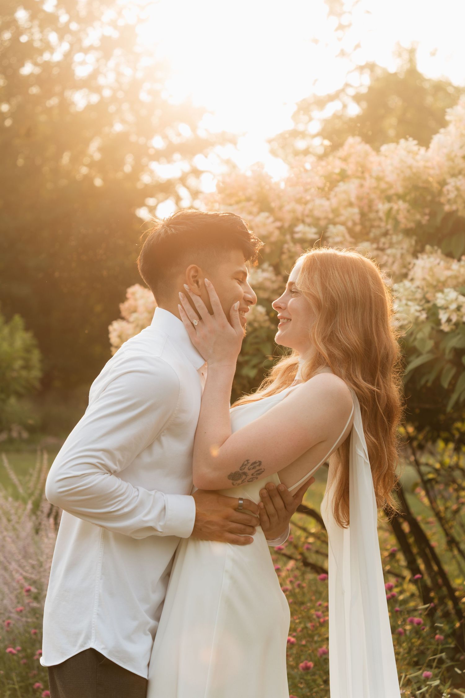 Couple sharing a romantic moment in a sunlit garden with blooming flowers, woman in white dress smiling as man leans in.