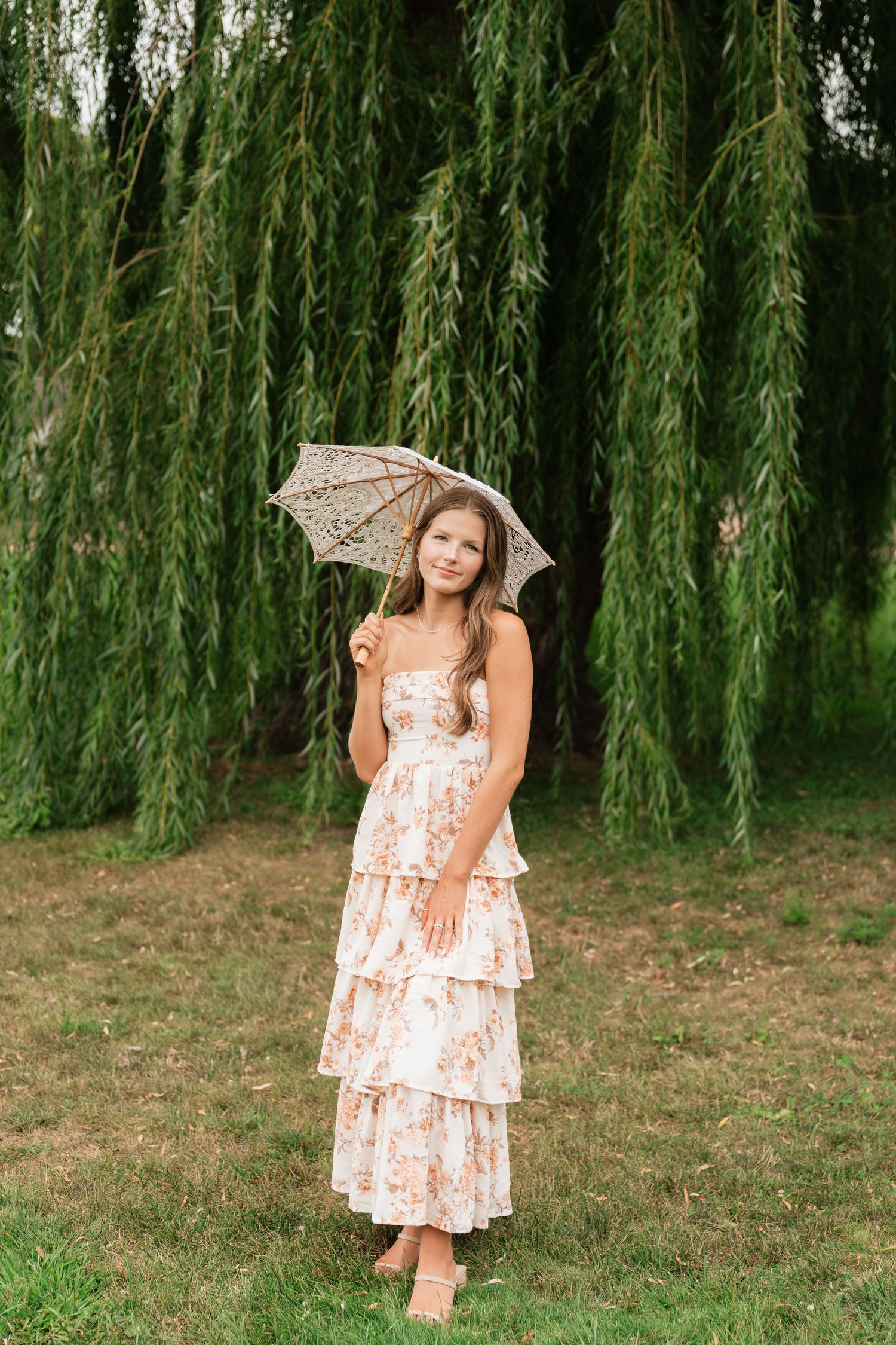 Young woman in floral tiered dress holding lace parasol, standing barefoot on grass before weeping willow tree.