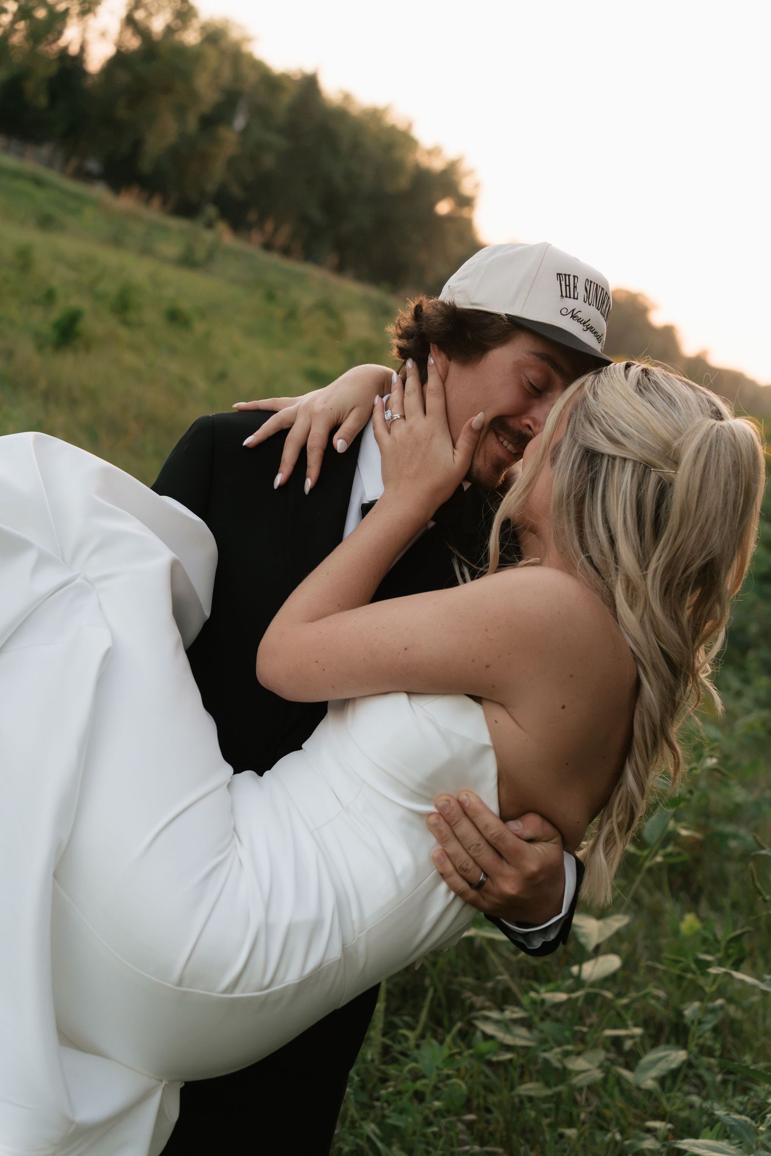 Groom in a baseball cap holds bride in white dress as they kiss in a lush green field at golden hour.