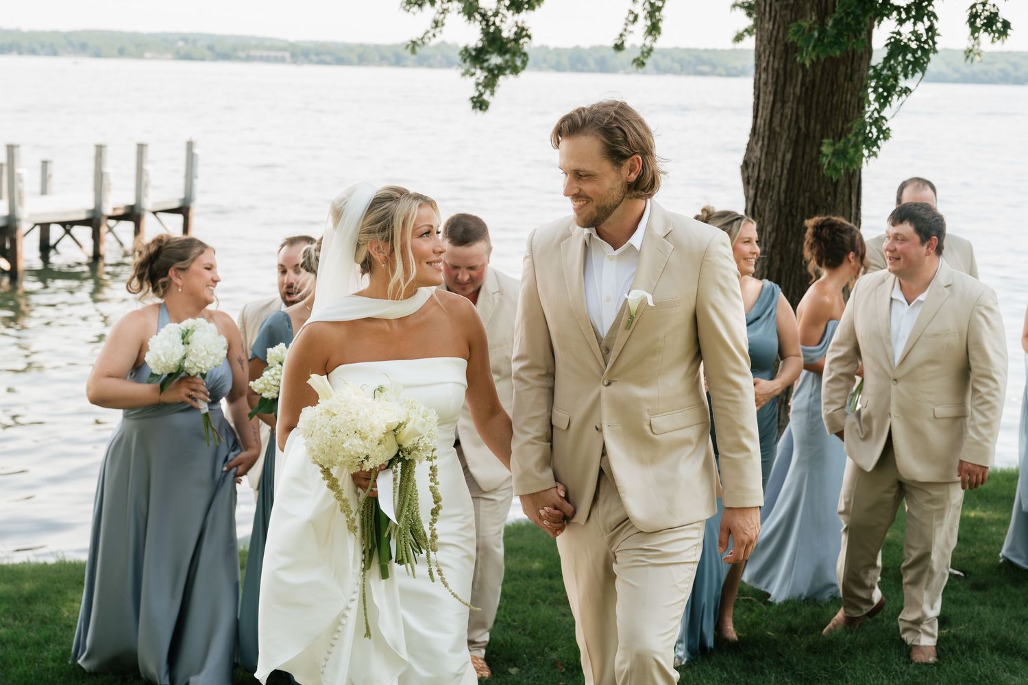 Bride and groom walk hand-in-hand past wedding party in blue gowns and tan suits near a lakeside dock.