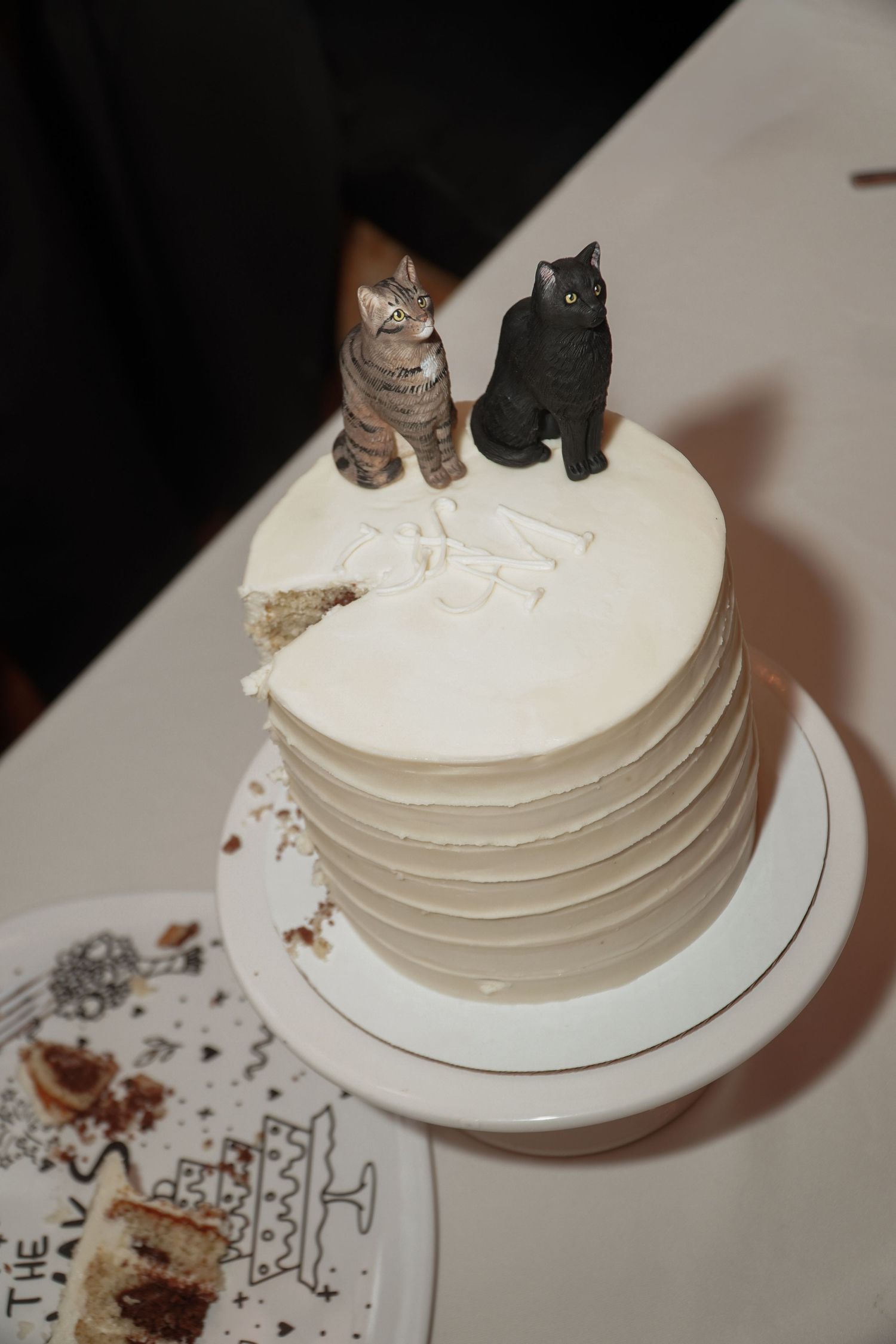 White wedding cake decorated with two cat figurines on top, one black and one brown, with textured horizontal lines.