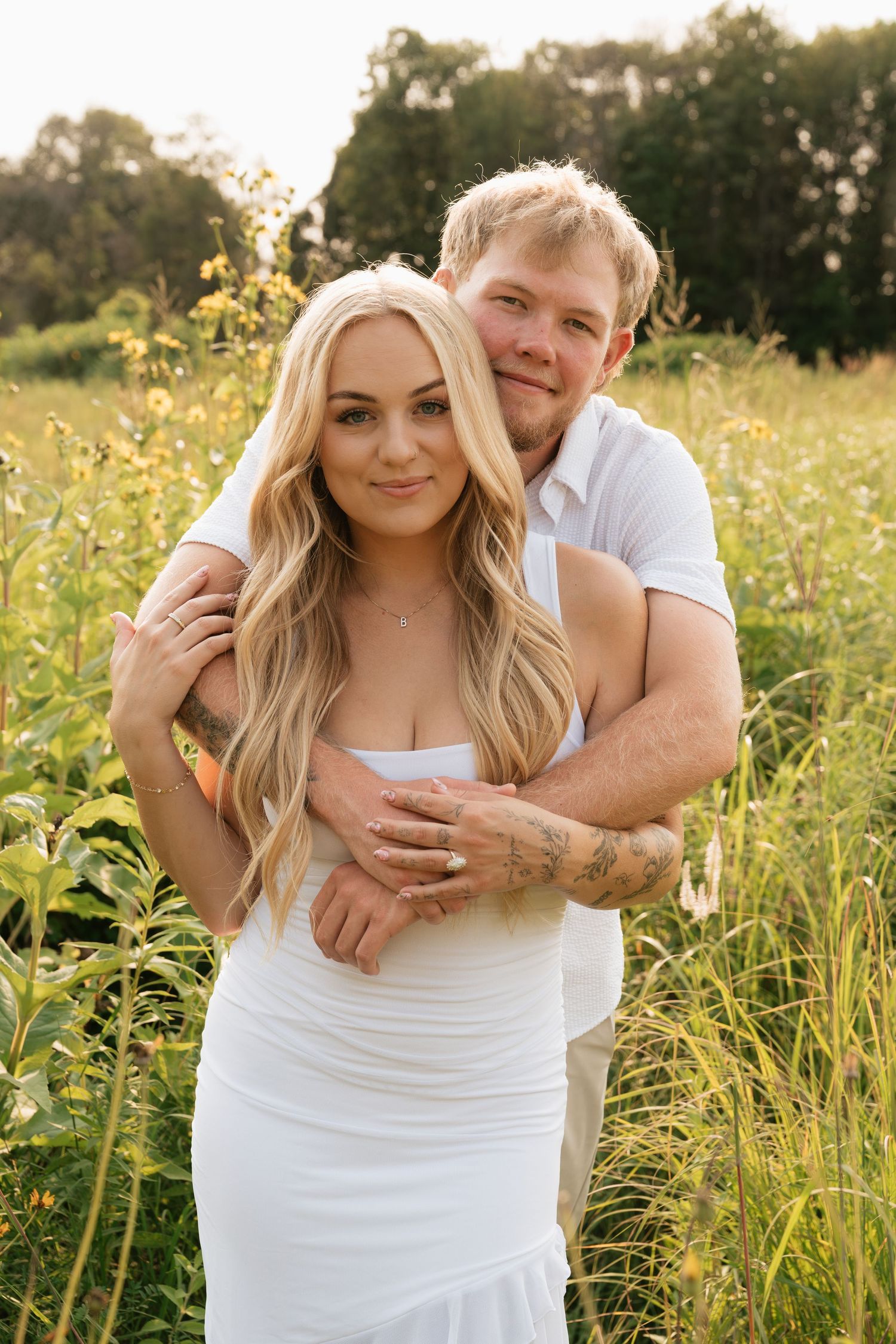 A couple embraces in a sunlit field, woman in white dress with long blonde hair, man in white shirt behind her.