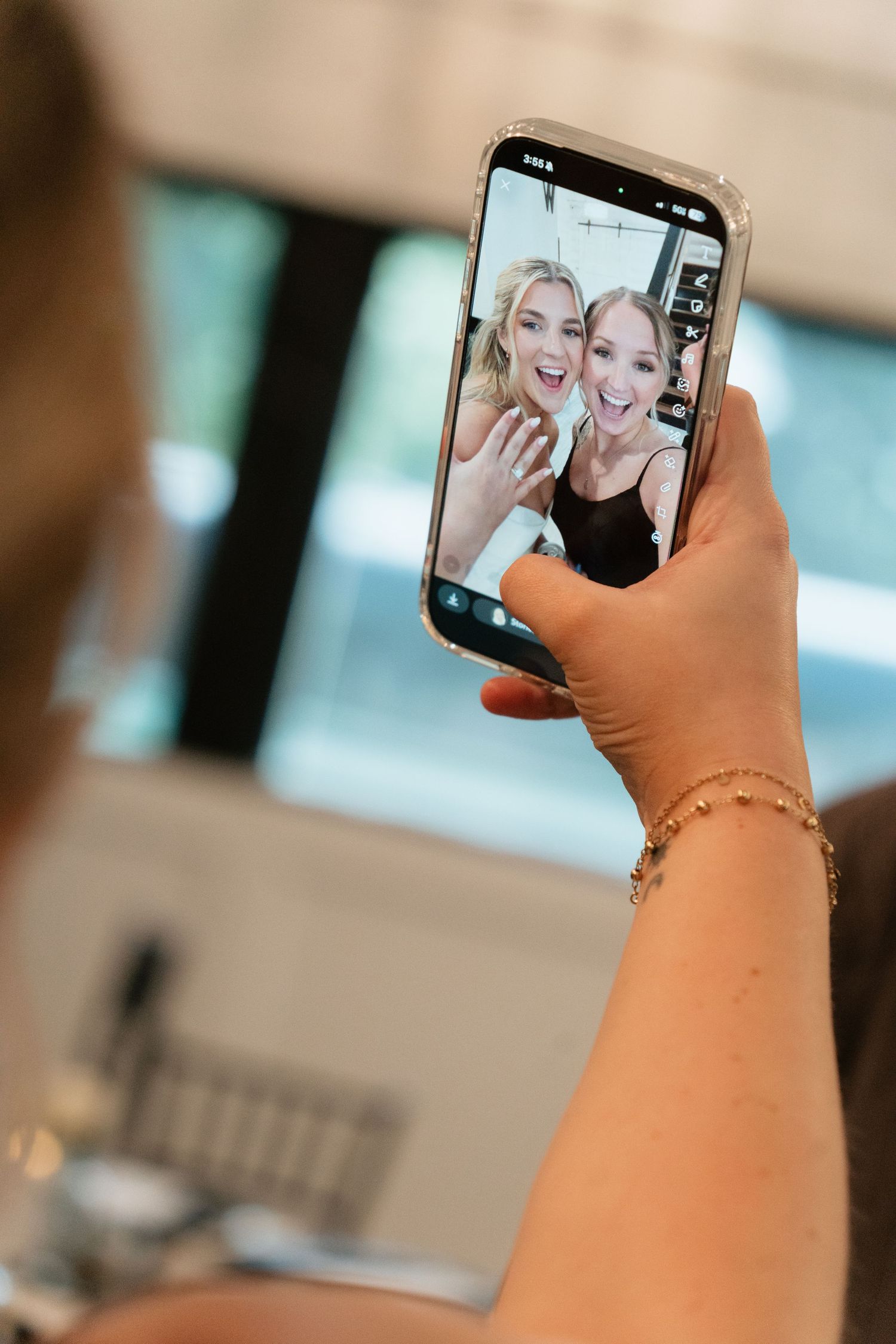 A hand holding a smartphone displaying two smiling women waving during a video call, with a gold bracelet visible.