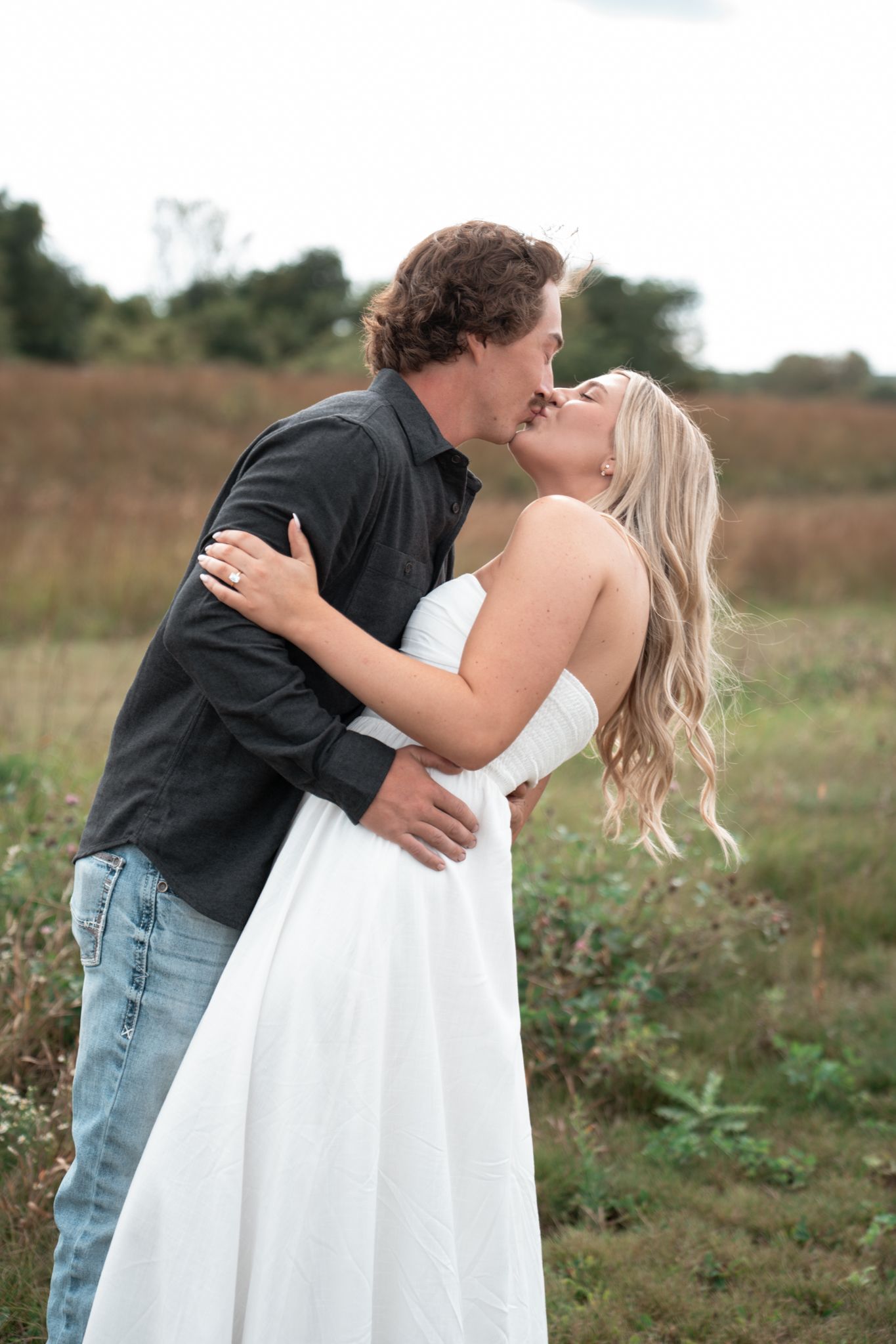 Couple embracing in a rustic outdoor field wearing formal attire.