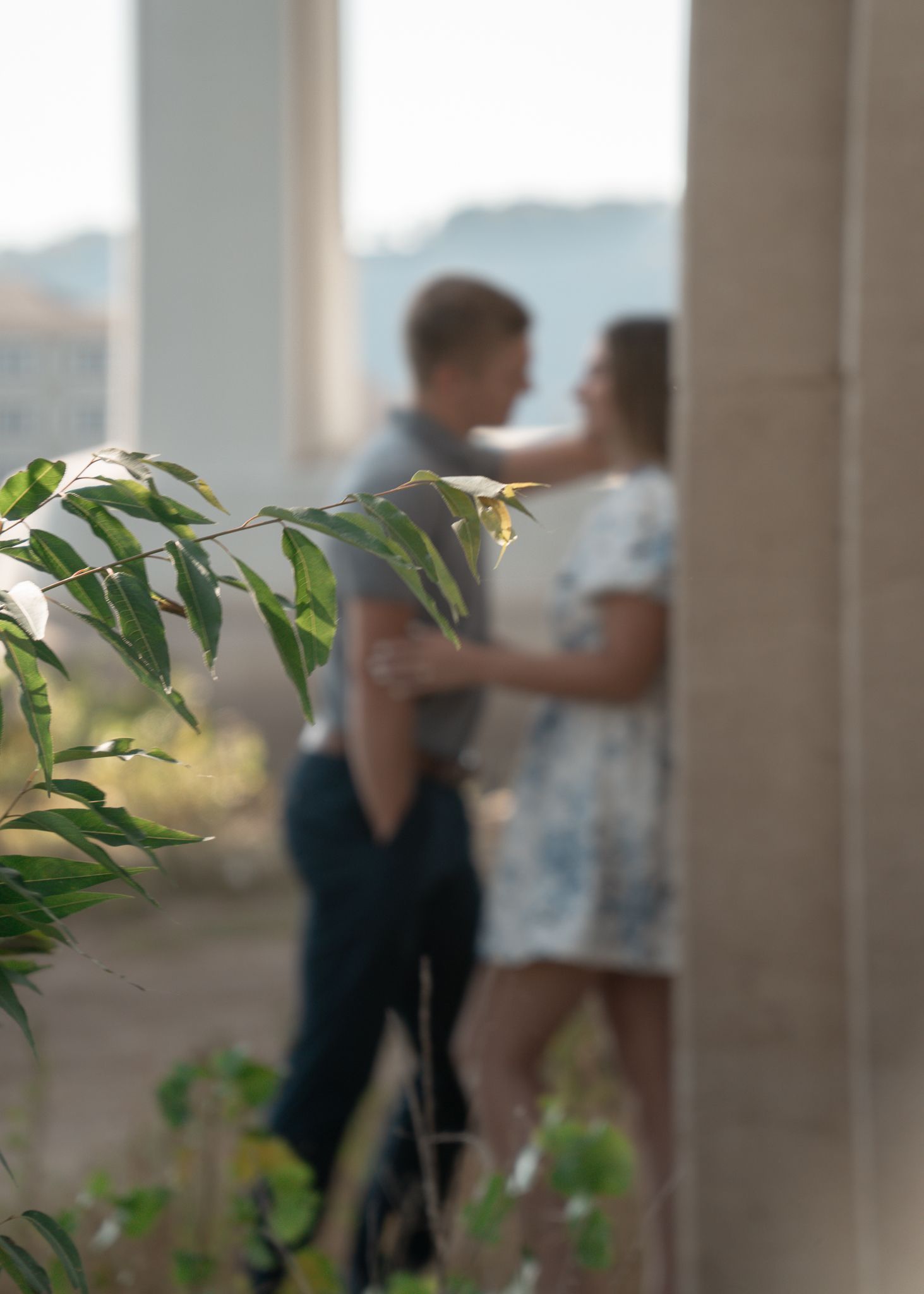 A romantic couple embraces on a balcony with city views and greenery in the background.