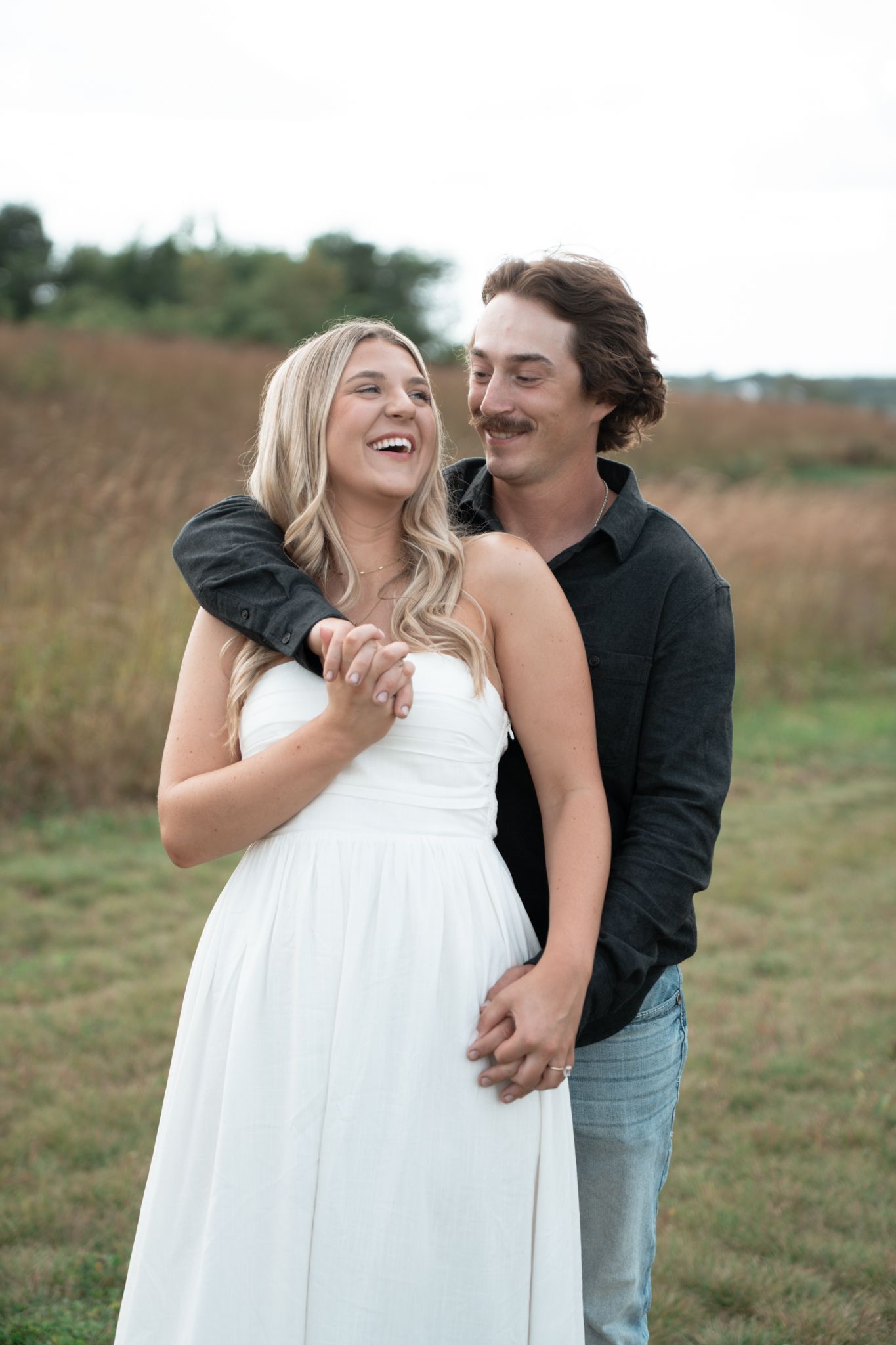 A couple shares a joyful moment together in a grassy field, the bride wearing a flowing white dress.