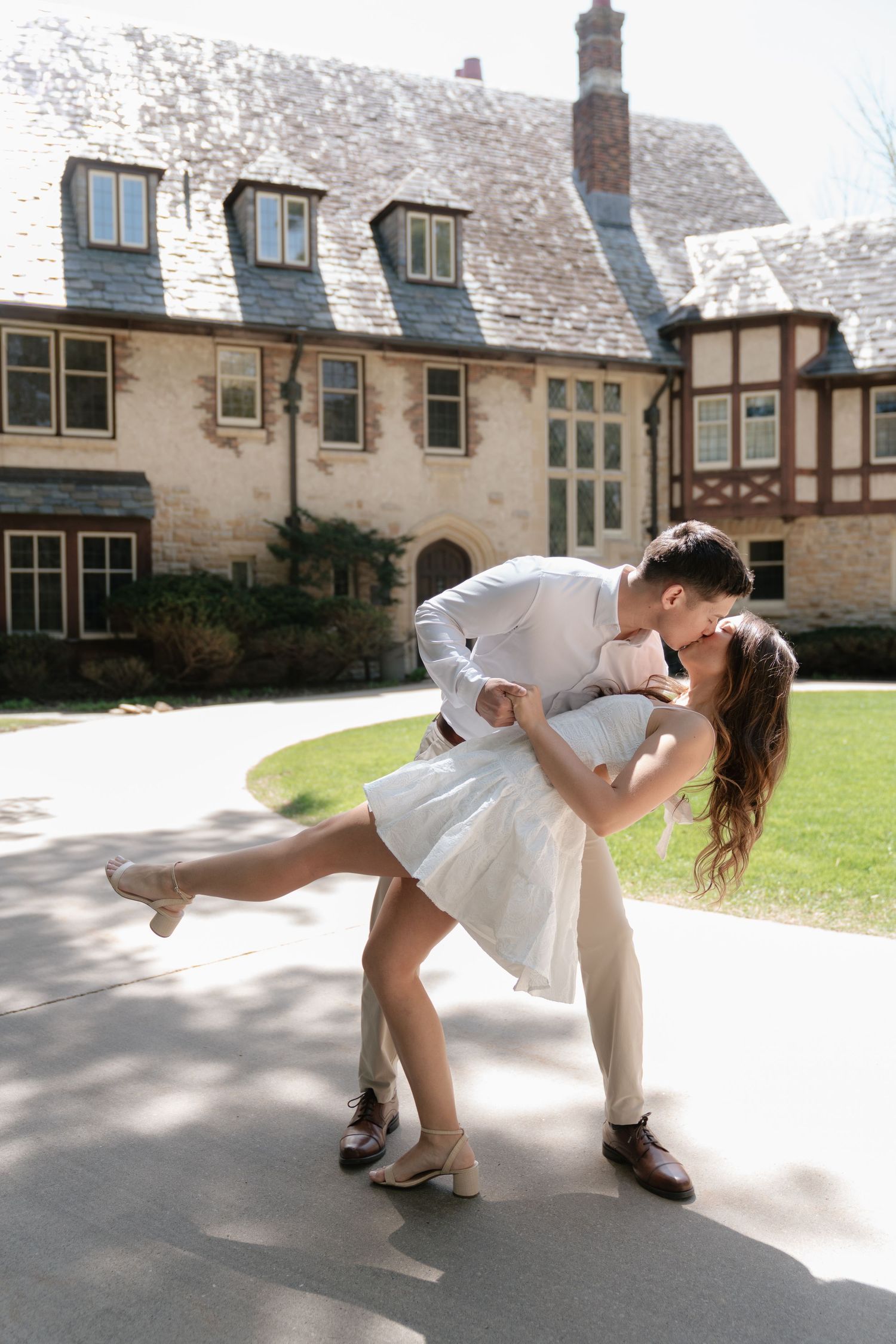 Couple sharing a romantic dip kiss in white outfits outside an elegant stone manor house on a sunny day.