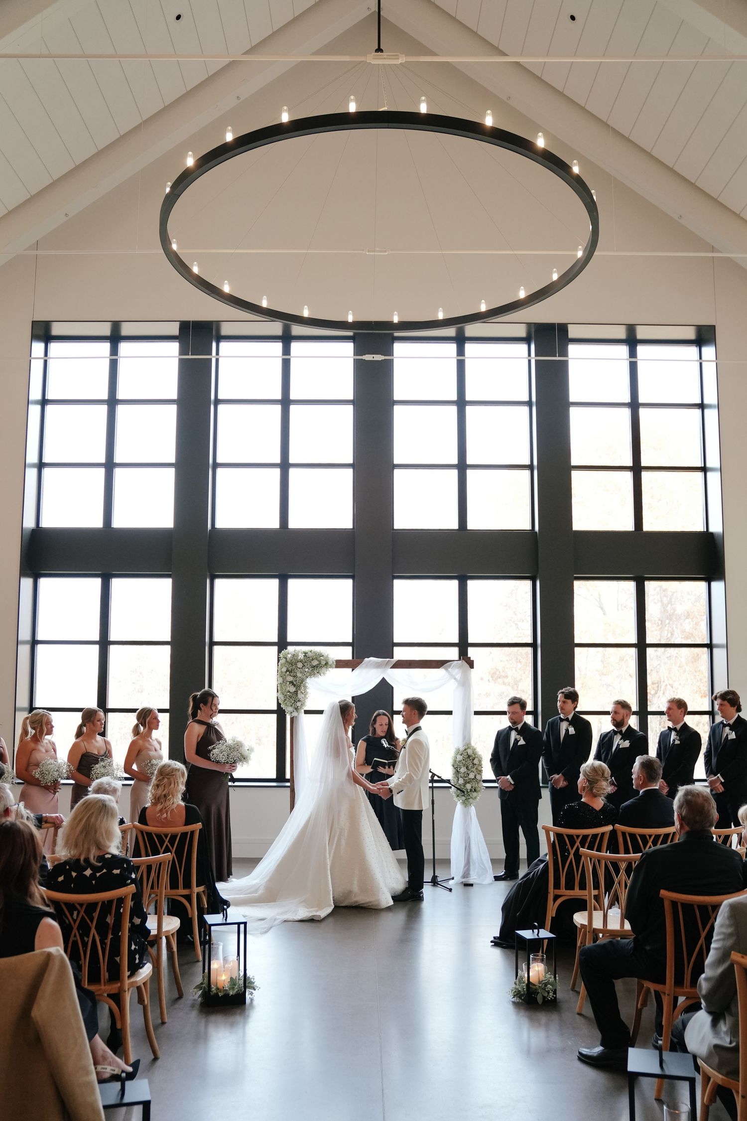 Indoor wedding ceremony in progress at a bright modern venue with white vaulted ceilings and large windows.