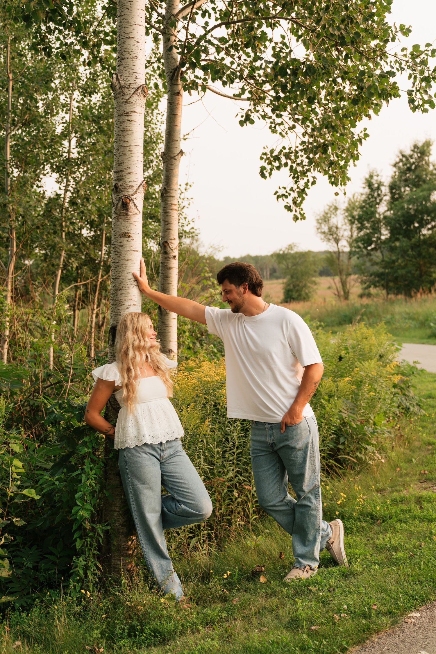 A couple in white tops and jeans leaning against a birch tree in a lush green outdoor setting.