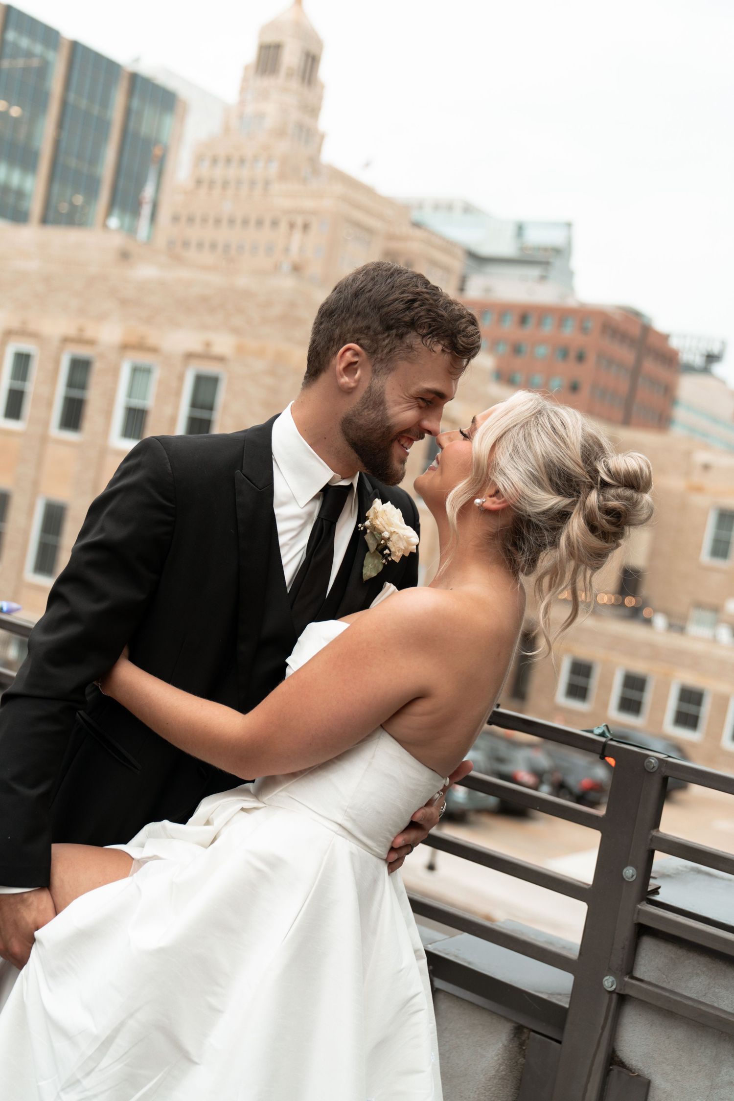 A bride and groom share a romantic moment on a rooftop balcony with a city skyline backdrop on their wedding day.
