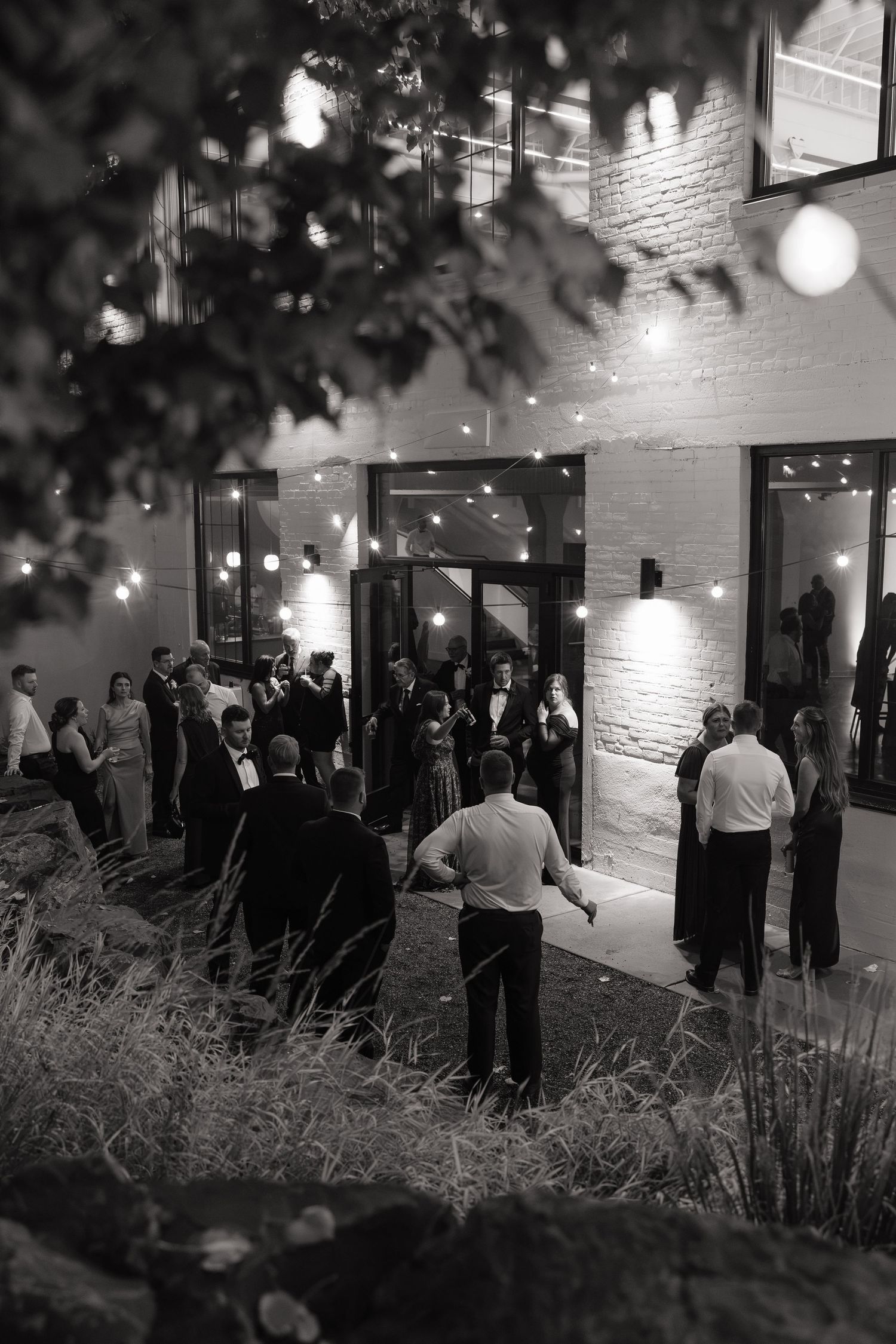 Black and white photo of elegantly dressed guests mingling outside a brick venue with string lights at night.