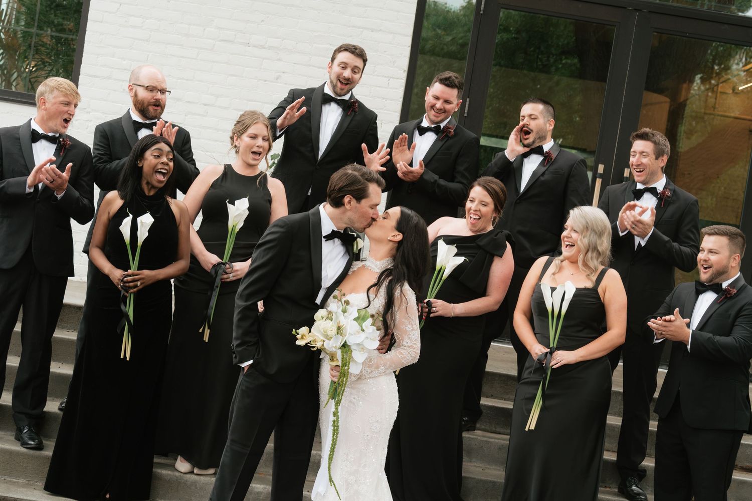 Wedding party in black attire cheers as bride and groom share a kiss on outdoor steps. The Lowlands wedding venue in St. Paul, MN.