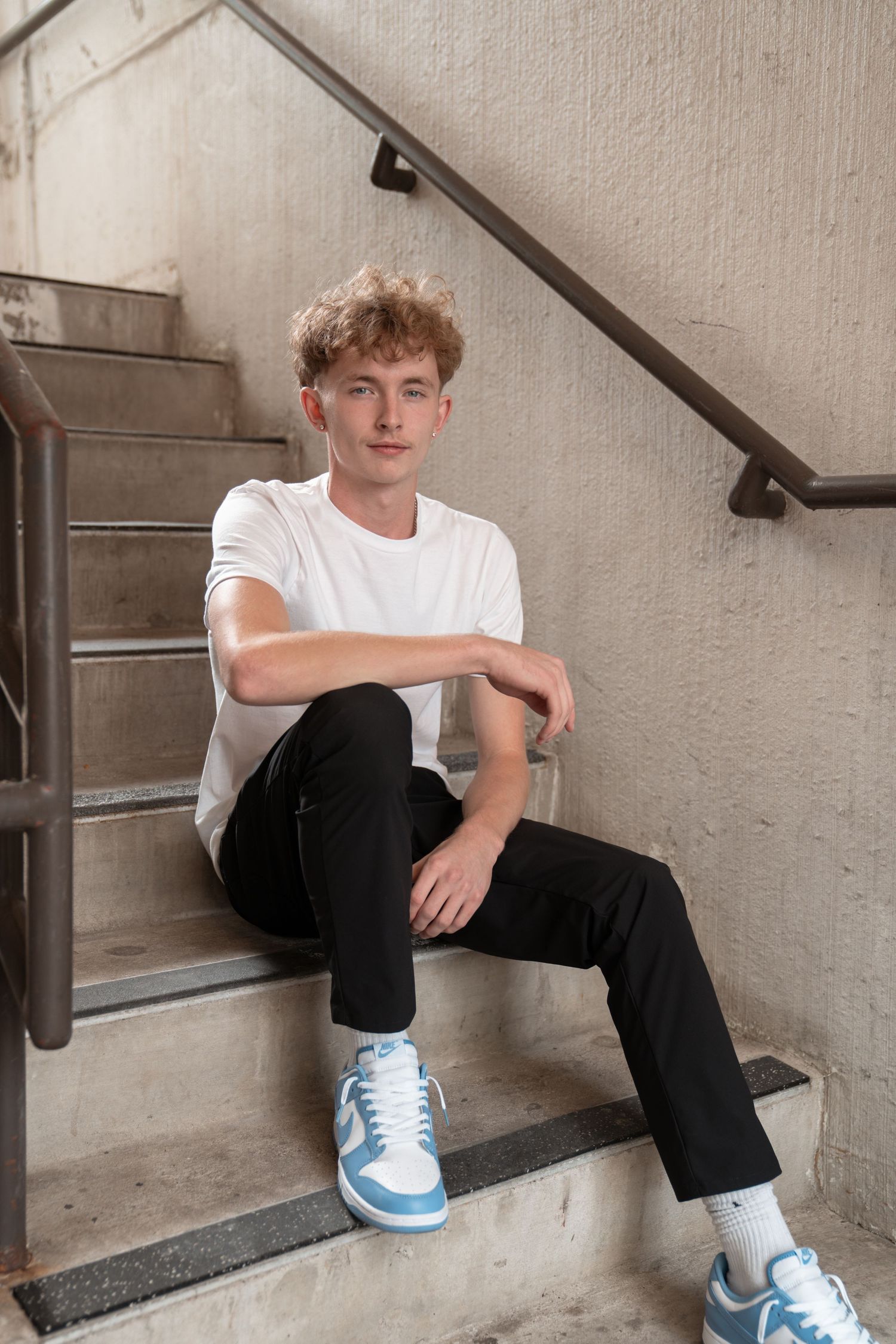 Person in white t-shirt and dark pants poses on concrete stairs wearing light blue sneakers.