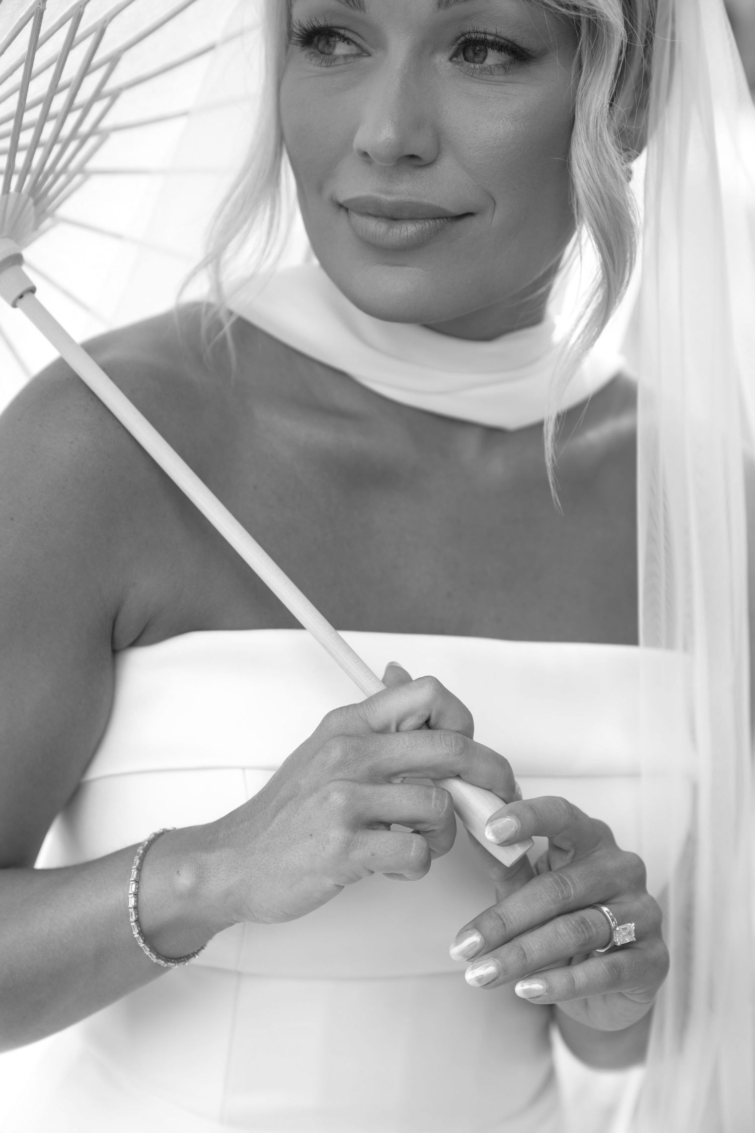 Black and white photo of a bride in white dress holding a parasol, wearing an engagement ring and bracelet.